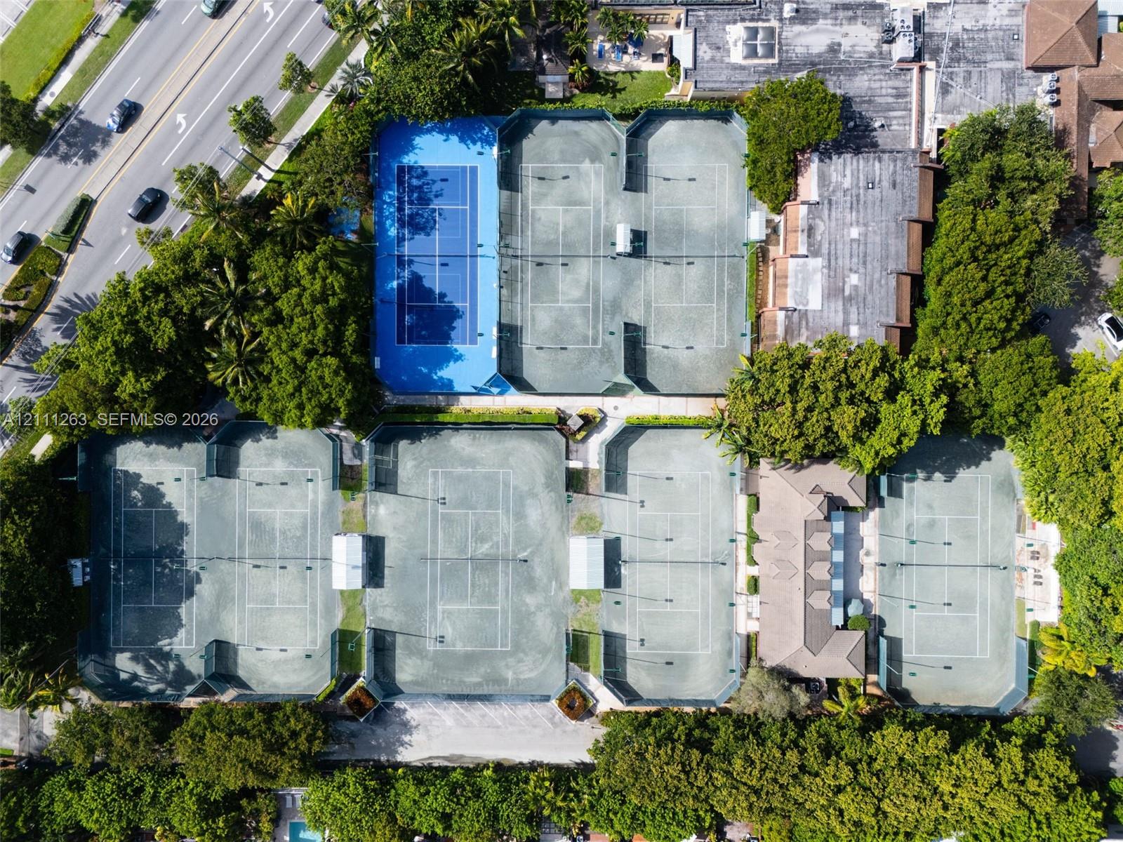 Aerial view of tennis complex featuring seven hard-court surfaces, perimeter fencing, and mature trees