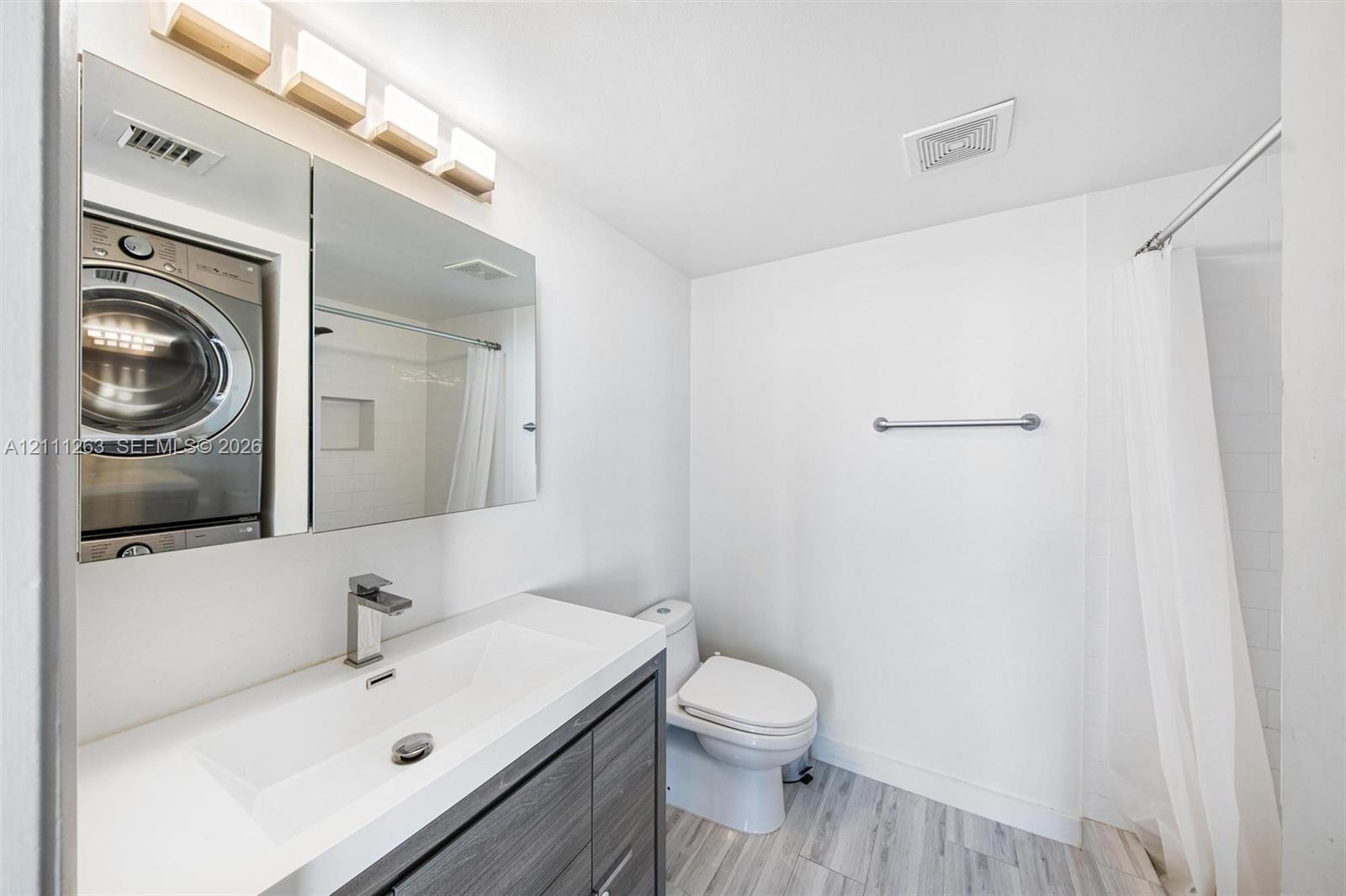 Wood-finish vanity with integrated rectangular sink and metallic faucet, mirrored medicine cabinet, stacked laundry appliance, and shower with tiled surround