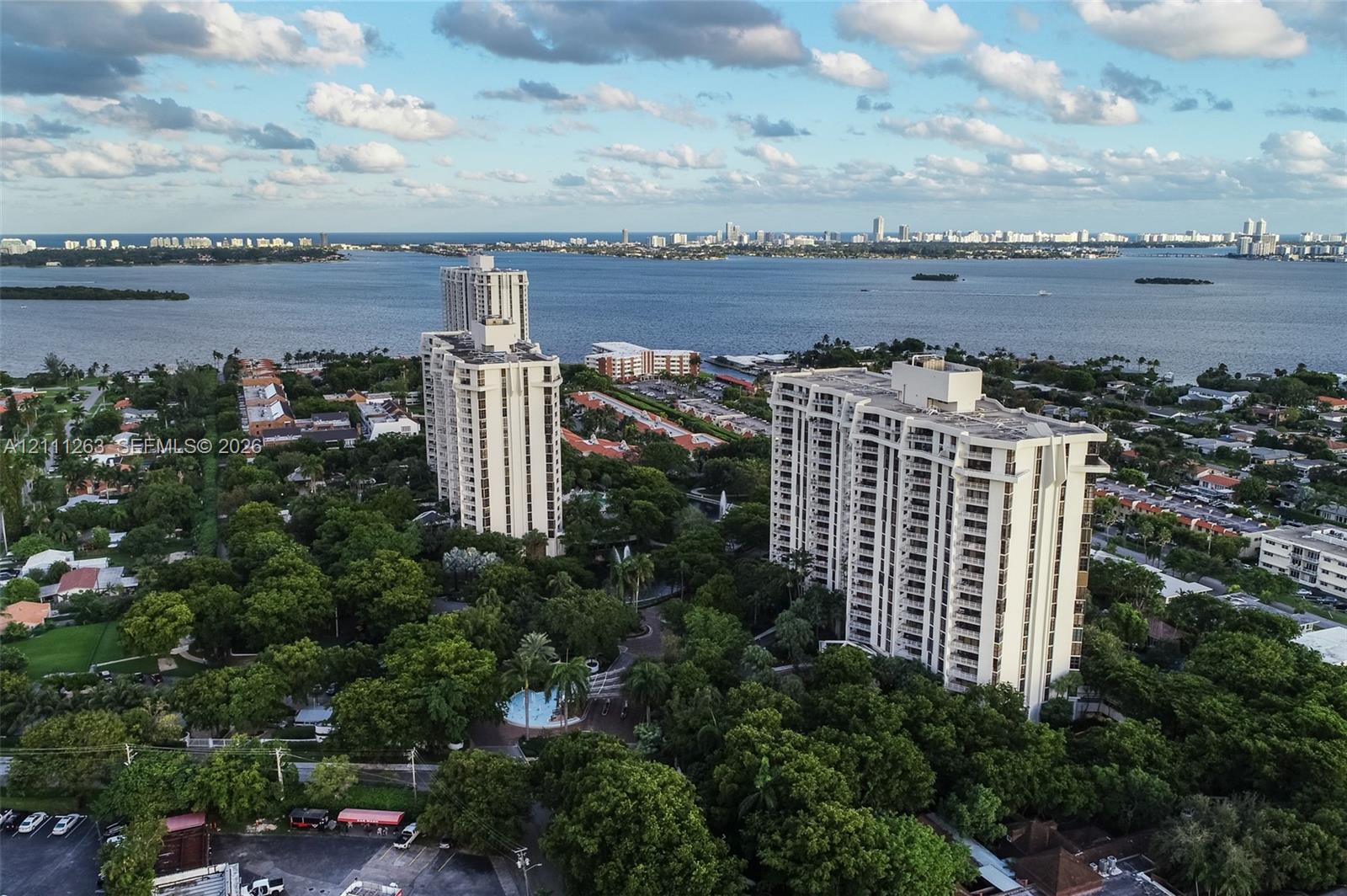 Aerial perspective featuring two high-rise towers, mature tree canopy, water backdrop, and circular swimming pool