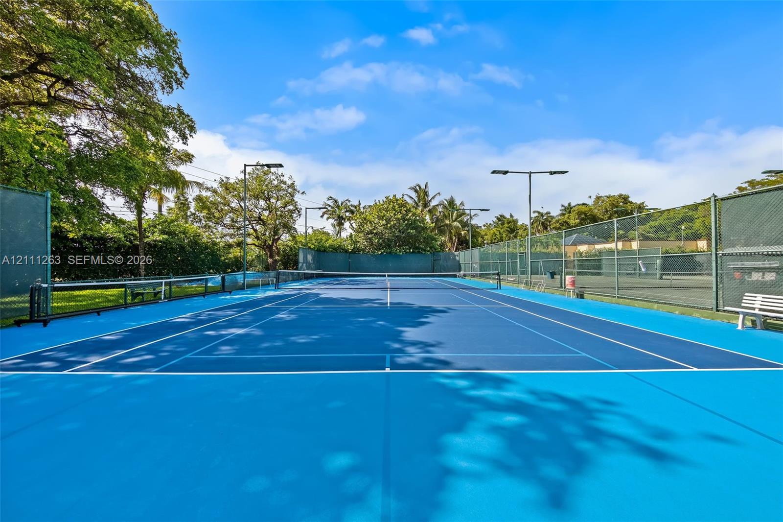 Hard-surface tennis court featuring perimeter chain-link fencing, overhead pole lighting, and built-in bench seating