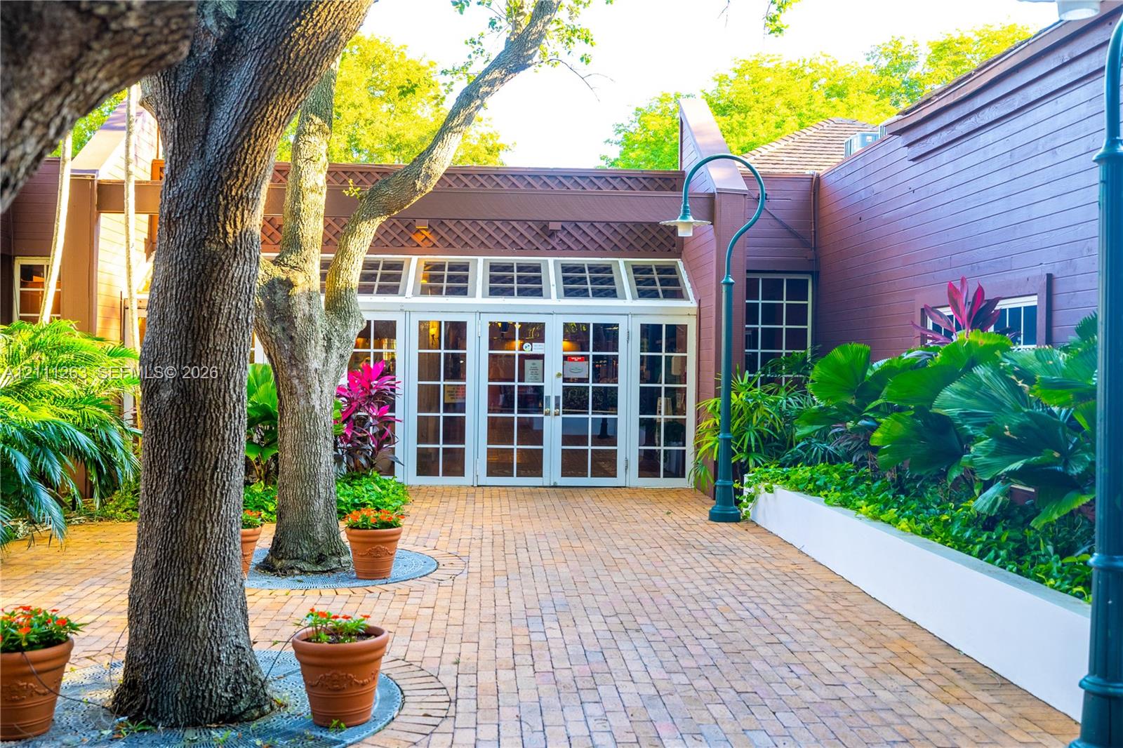 Exterior entrance featuring a brick-paved courtyard, wood-paneled siding, a multi-pane glass double-door entryway, and mature landscape plantings