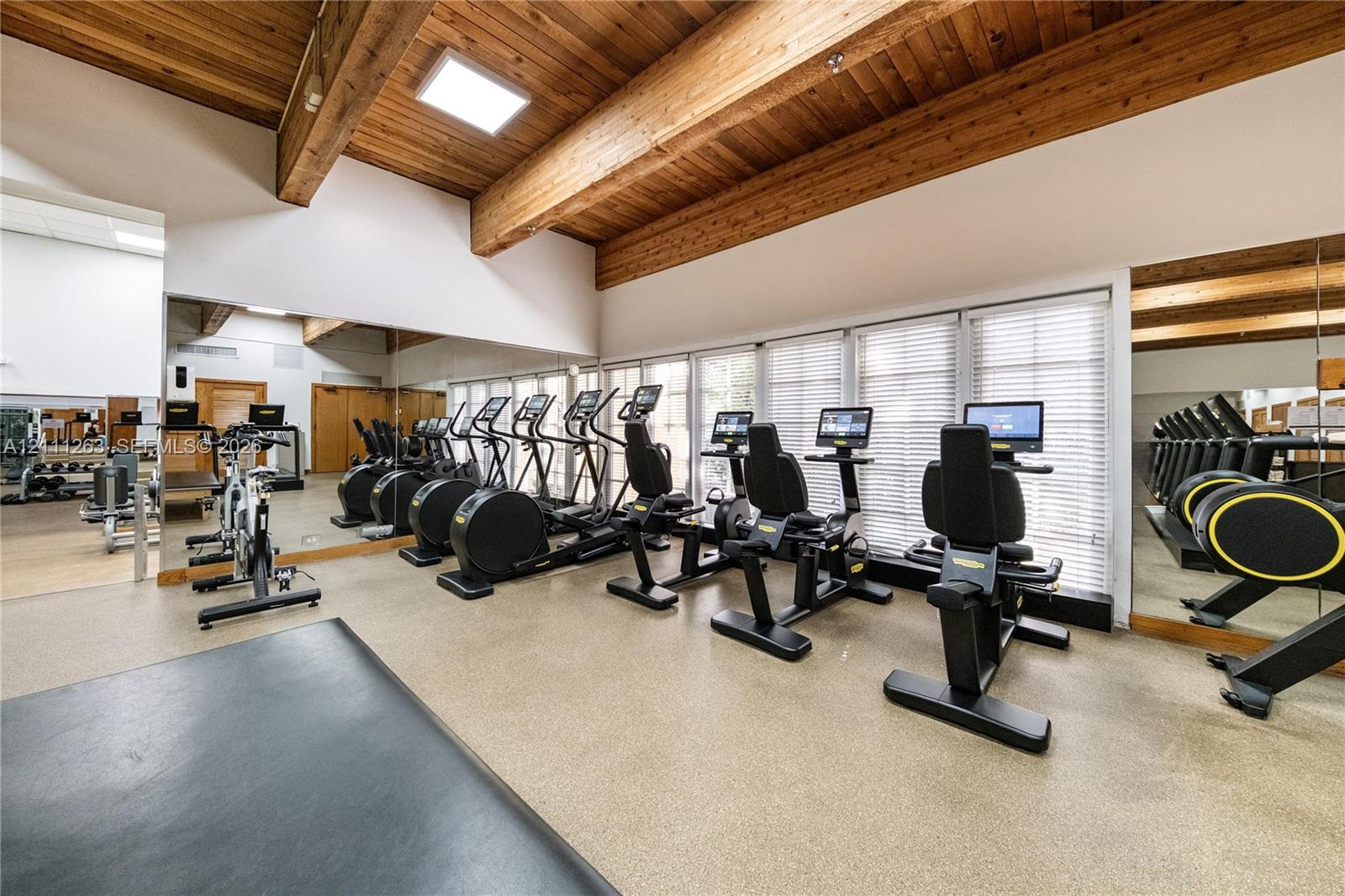 Fitness room featuring vaulted wood-paneled ceilings, exposed structural beams, floor-to-ceiling wall mirrors, and large windows with integrated blinds