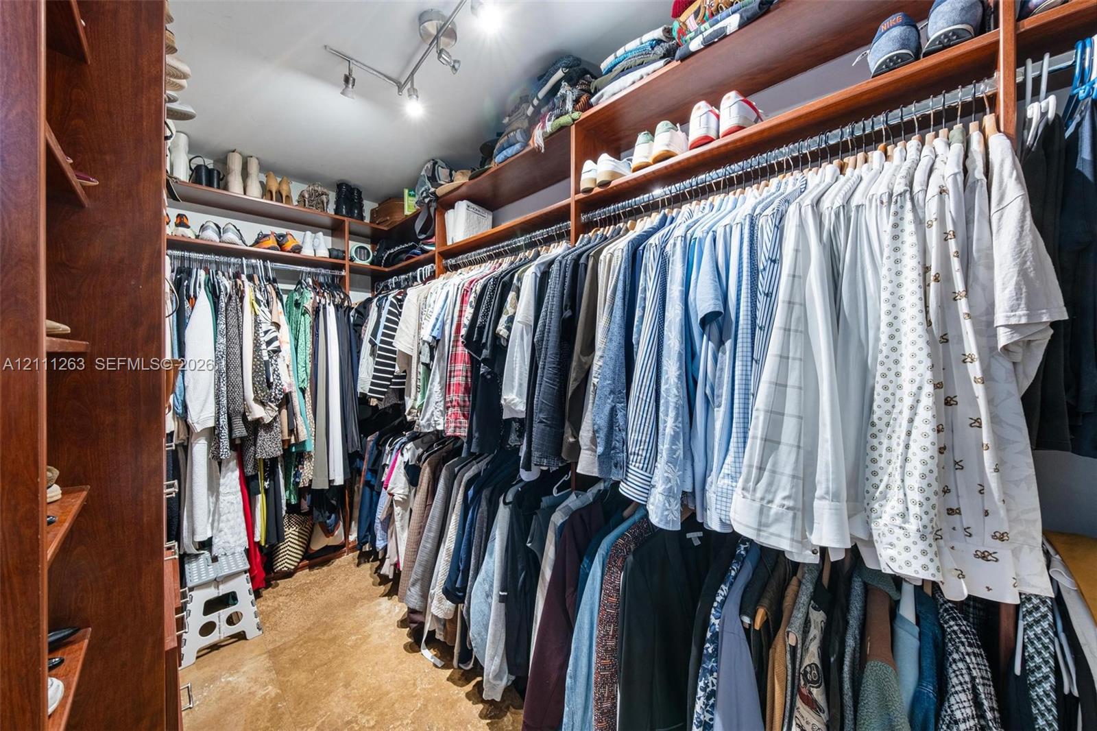 Walk-in closet featuring extensive wall-to-wall wood-finish cabinetry, tiered hanging rods, multiple open storage shelves, and ceiling-mounted directional track lighting