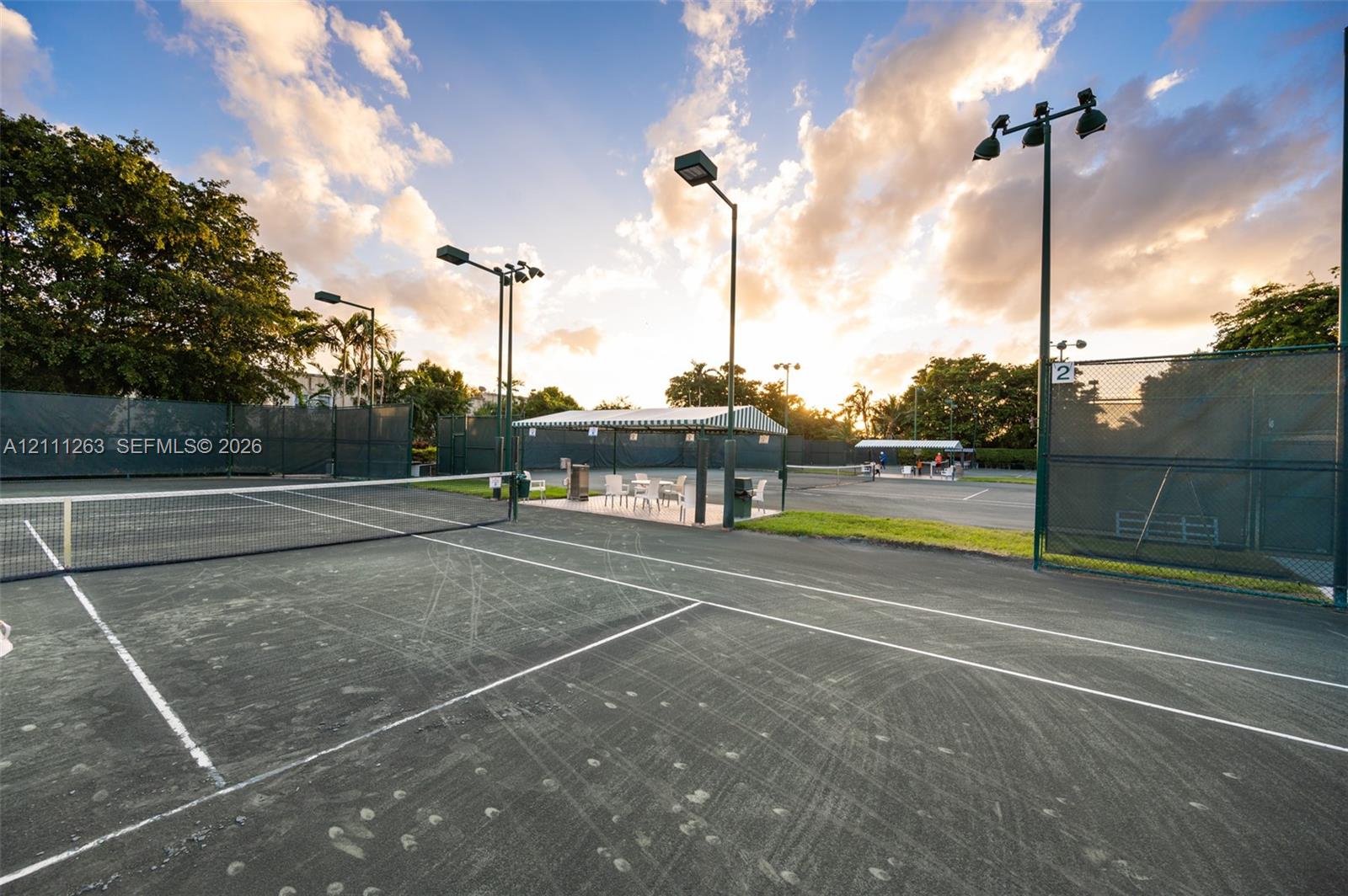 Hard-surfaced tennis court featuring perimeter fencing, overhead lighting fixtures, and a covered pavilion