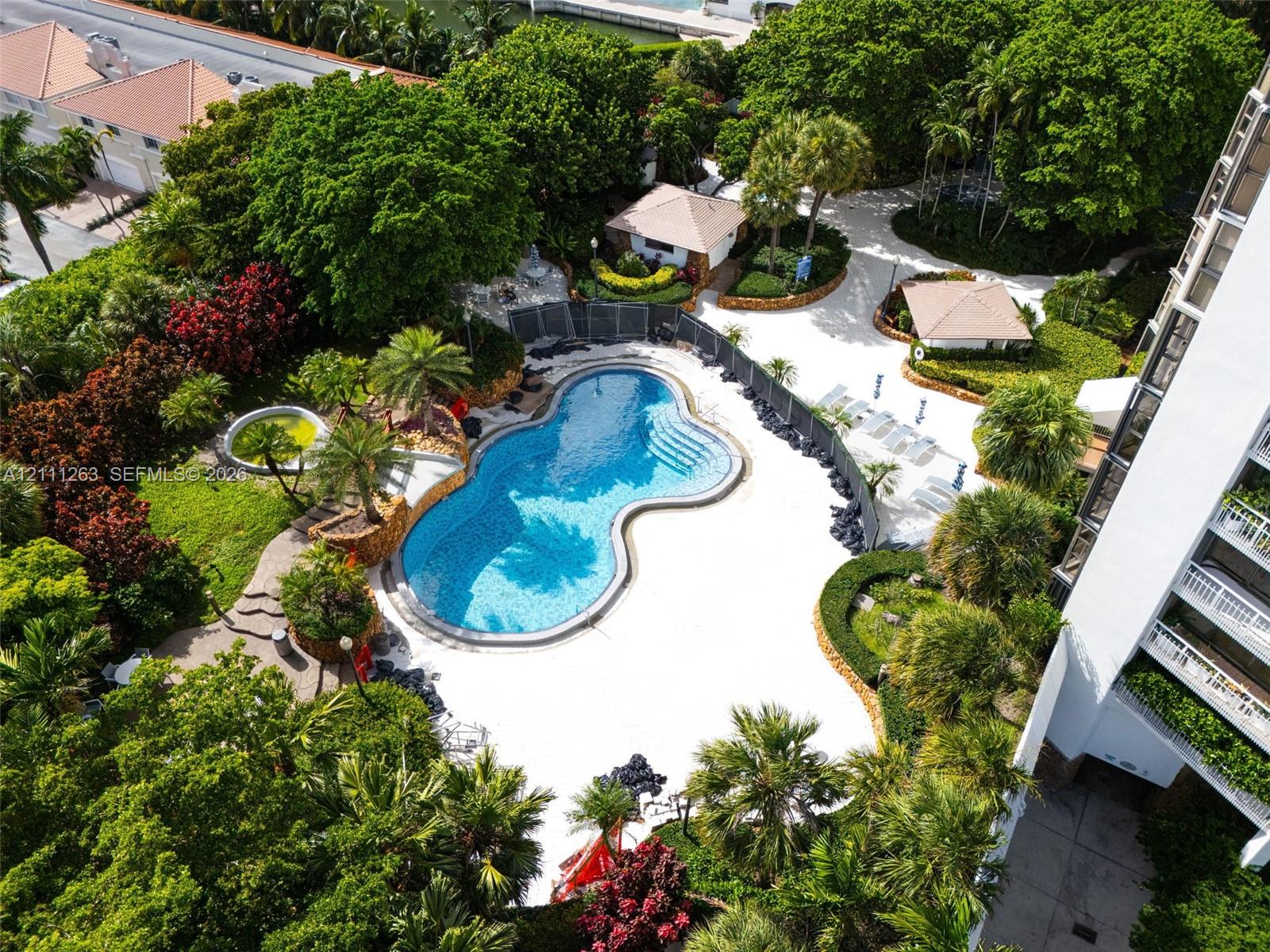 Freeform swimming pool with stone-accented perimeter, expansive paved sundeck, mature tropical landscaping, and dedicated covered structures