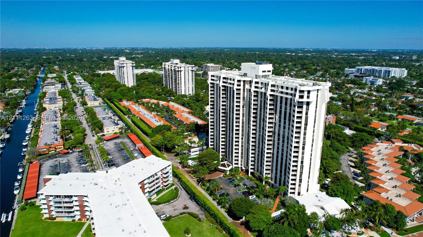 Aerial perspective of a high-rise residential structure featuring tiered balconies, covered parking areas, manicured grounds, and immediate access to a waterway
