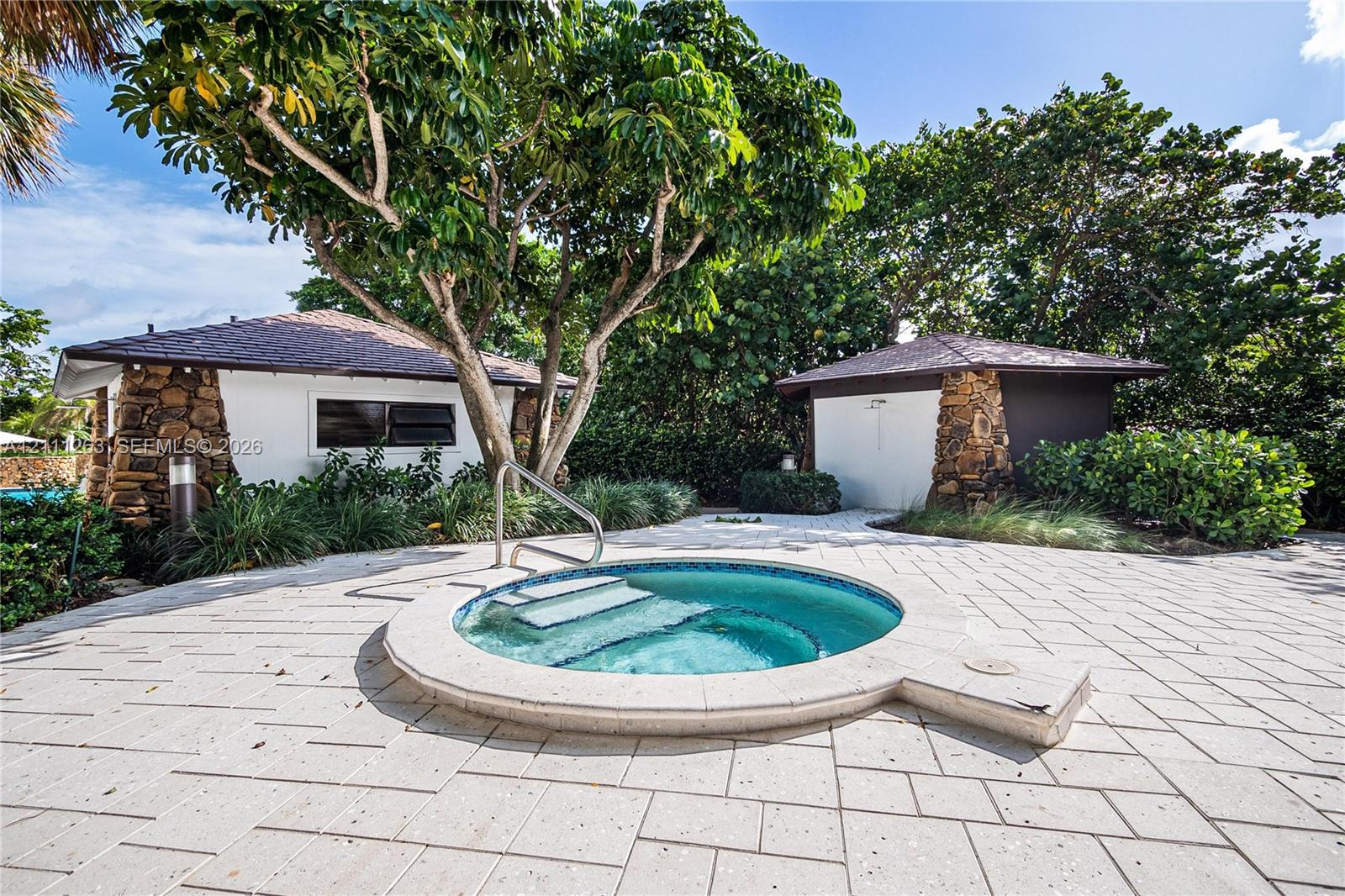 In-ground spa featuring a circular concrete surround, stone-accented exterior structures, and light-toned paver hardscaping