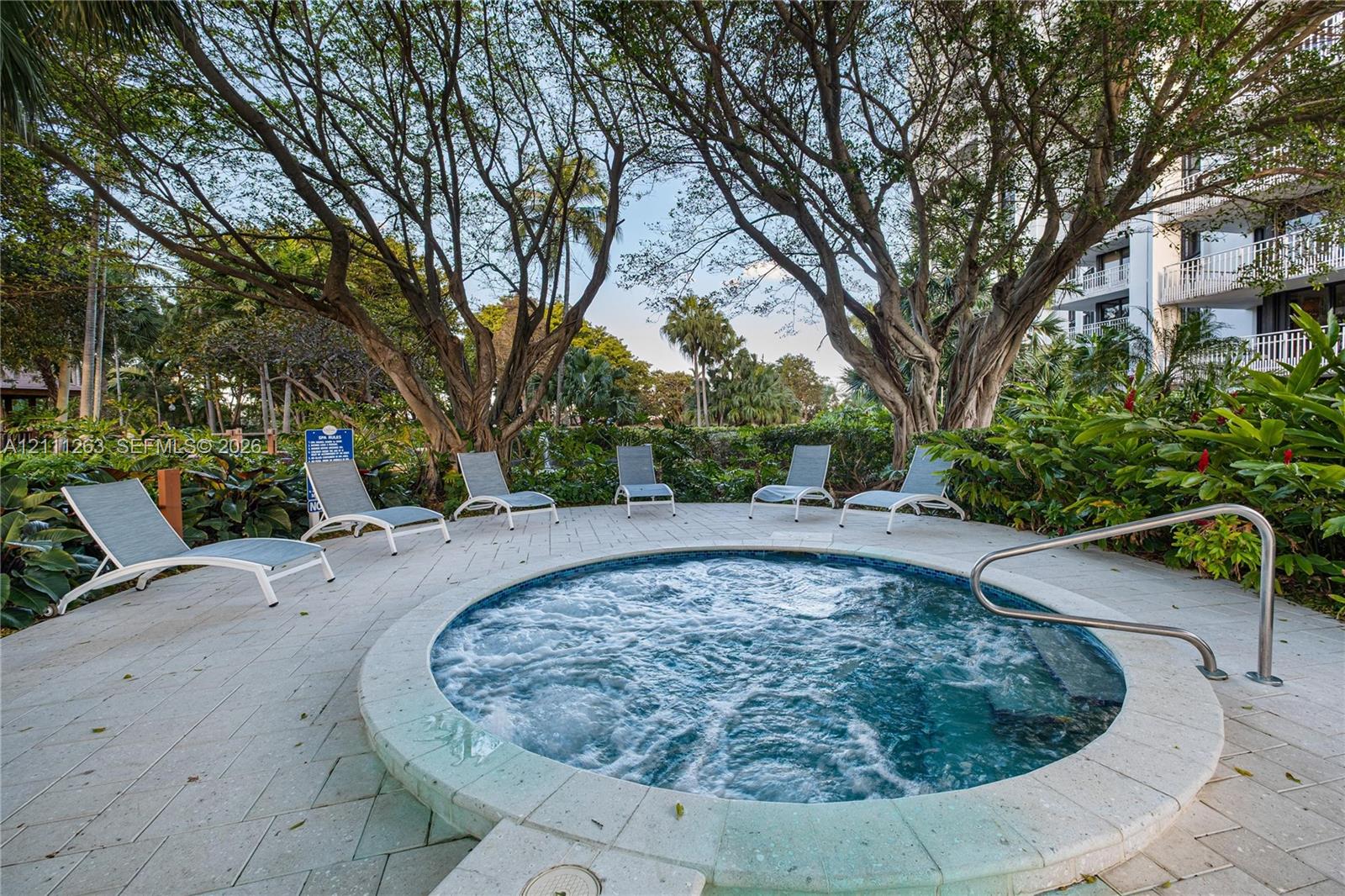 In-ground circular spa featuring a light-colored paver surround, integrated metal handrail, and mature surrounding landscaping