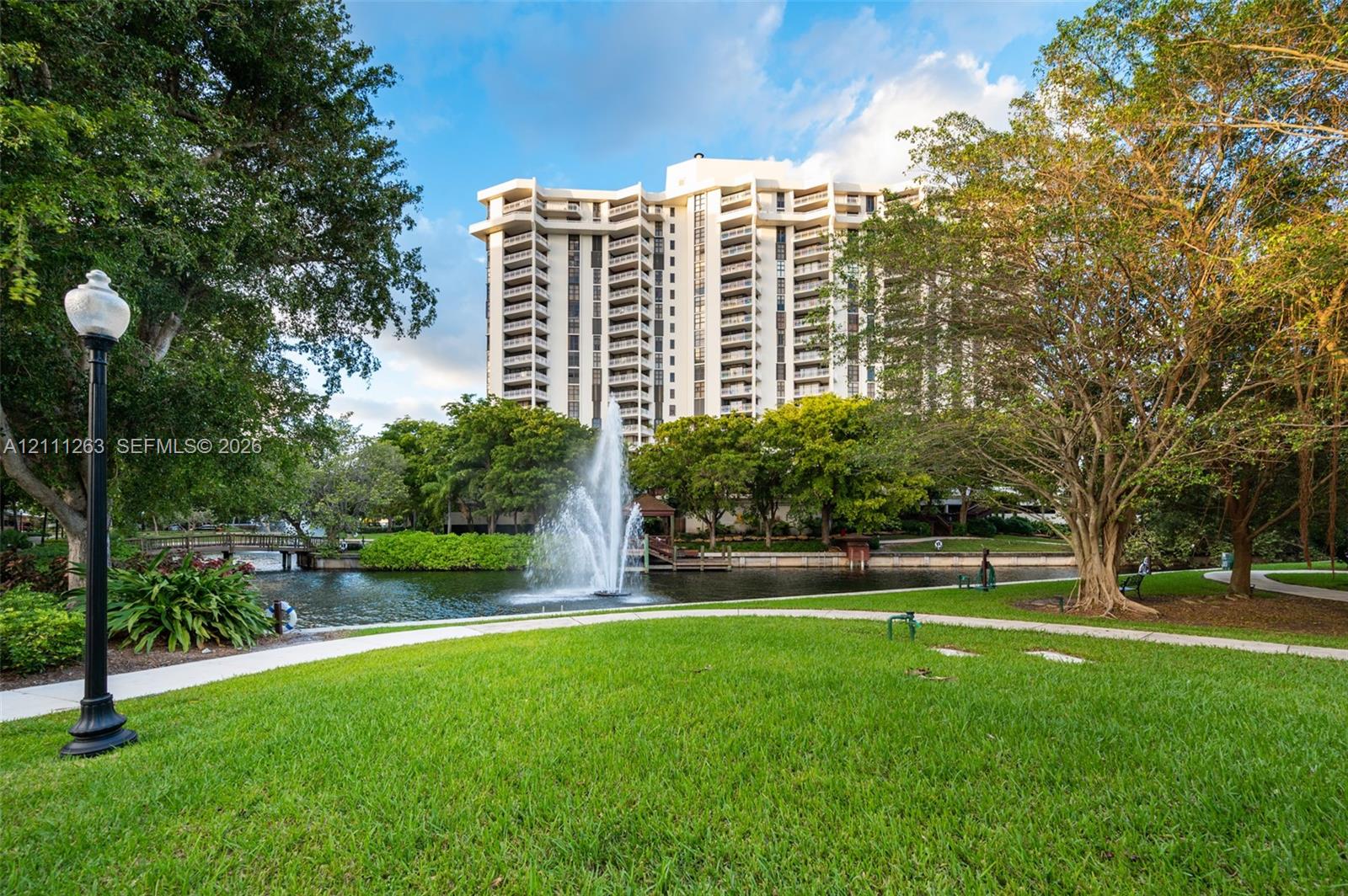 High-rise residential exterior featuring private lake frontage, a central water fountain, landscaped grounds, and paved walking paths