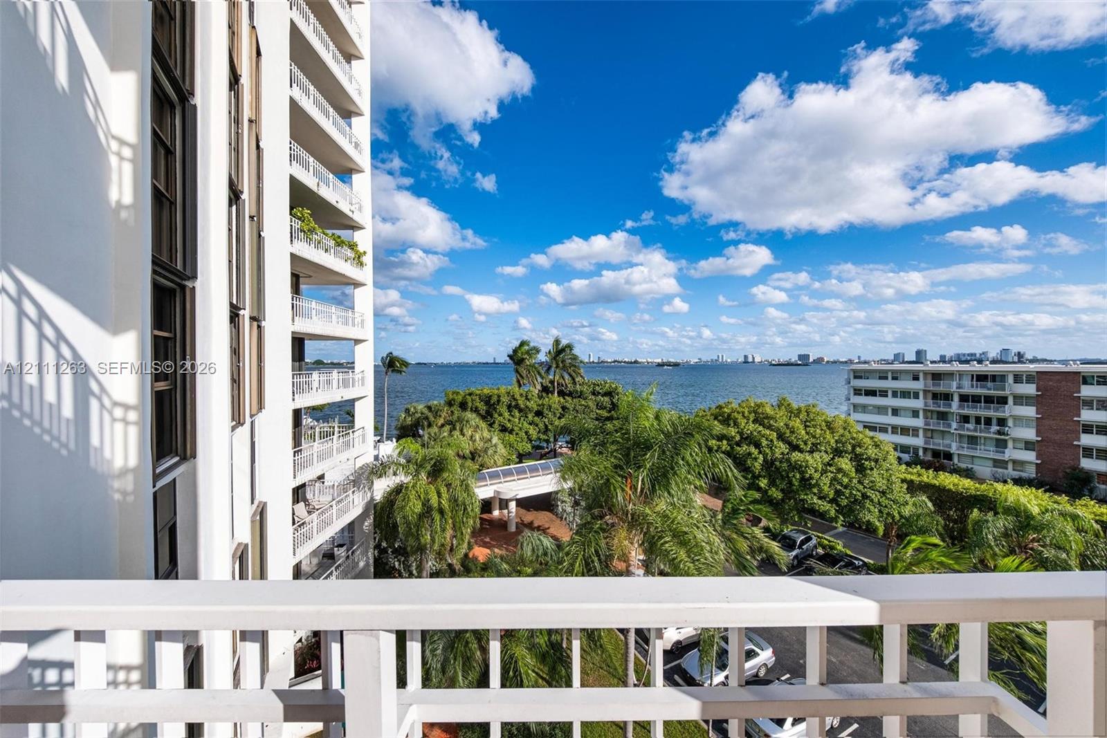 Private balcony with white railing, featuring expansive water scenery, tropical landscaping, and mature palm trees