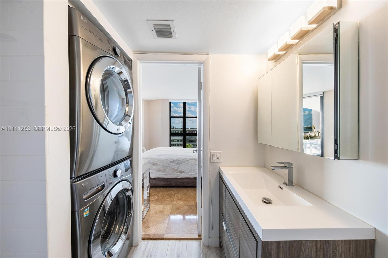 Stacked front-loading laundry appliances, built-in vanity with integrated rectangular sink, quartz countertop, multi-panel mirrored medicine cabinet, and contemporary single-handle faucet