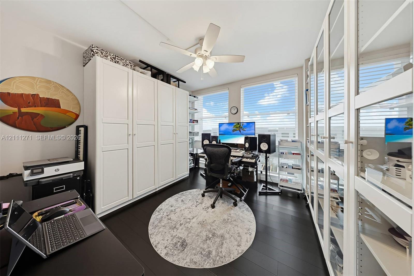 Custom built-in white cabinetry, expansive glass-paned storage units, dark wood-finish flooring, and a multi-blade ceiling fan with integrated lighting