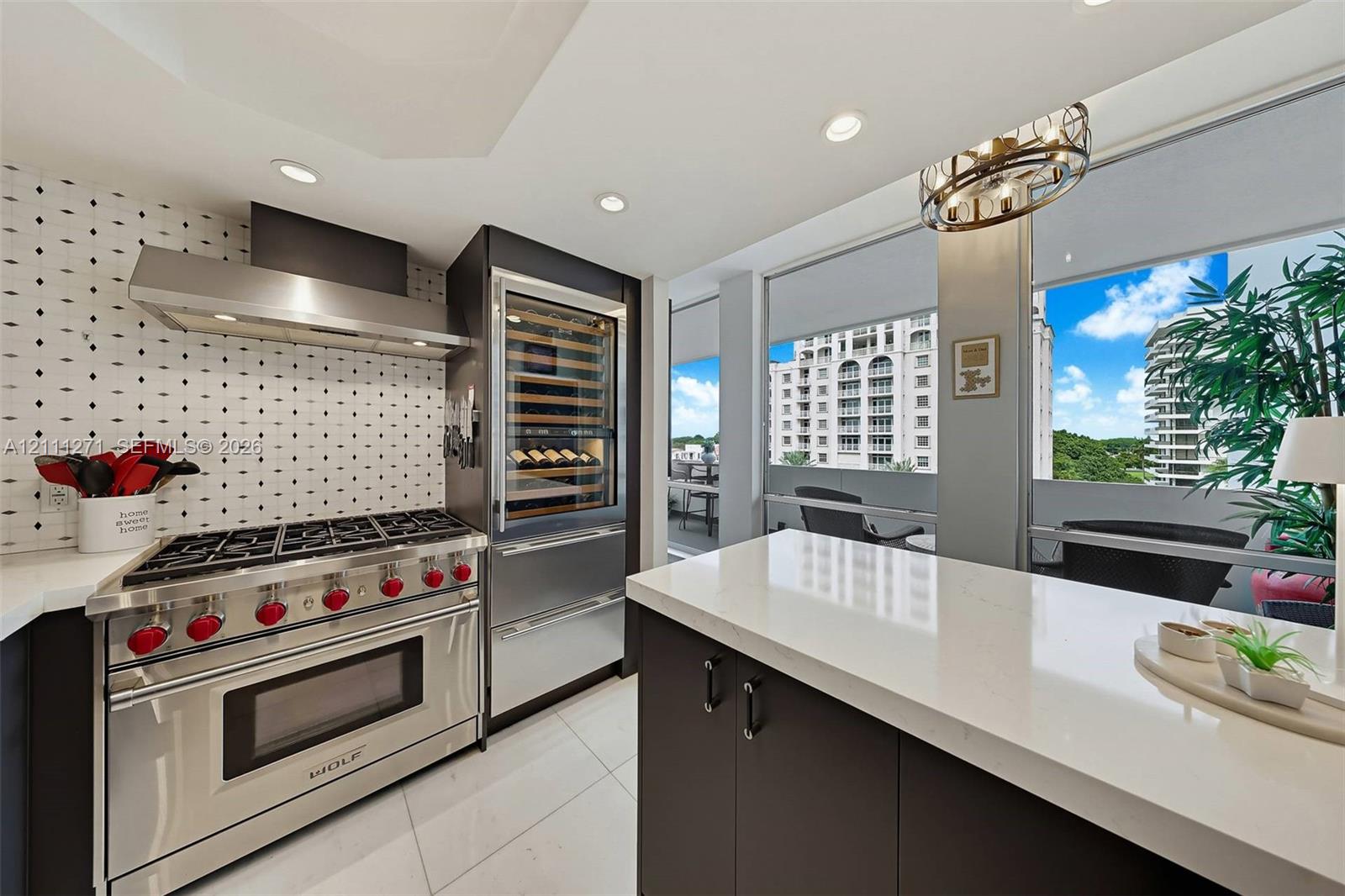 Kitchen featuring a stainless steel Wolf gas range, integrated wine refrigeration, stone-surfaced island, patterned tile backsplash, and floor-to-ceiling windows