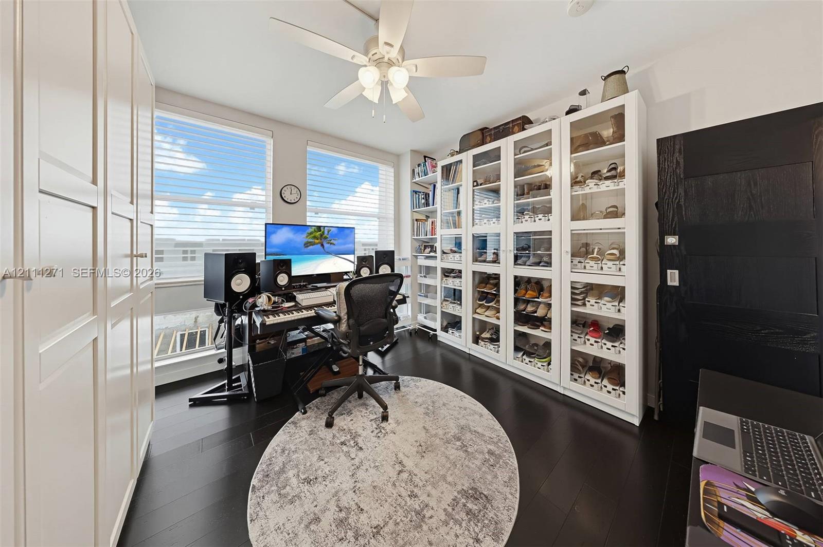Room featuring dark-stained wood-finish flooring, floor-to-ceiling built-in white cabinetry with glass-front doors, tall windows with horizontal blinds, and a multi-blade ceiling fan with integrated lighting