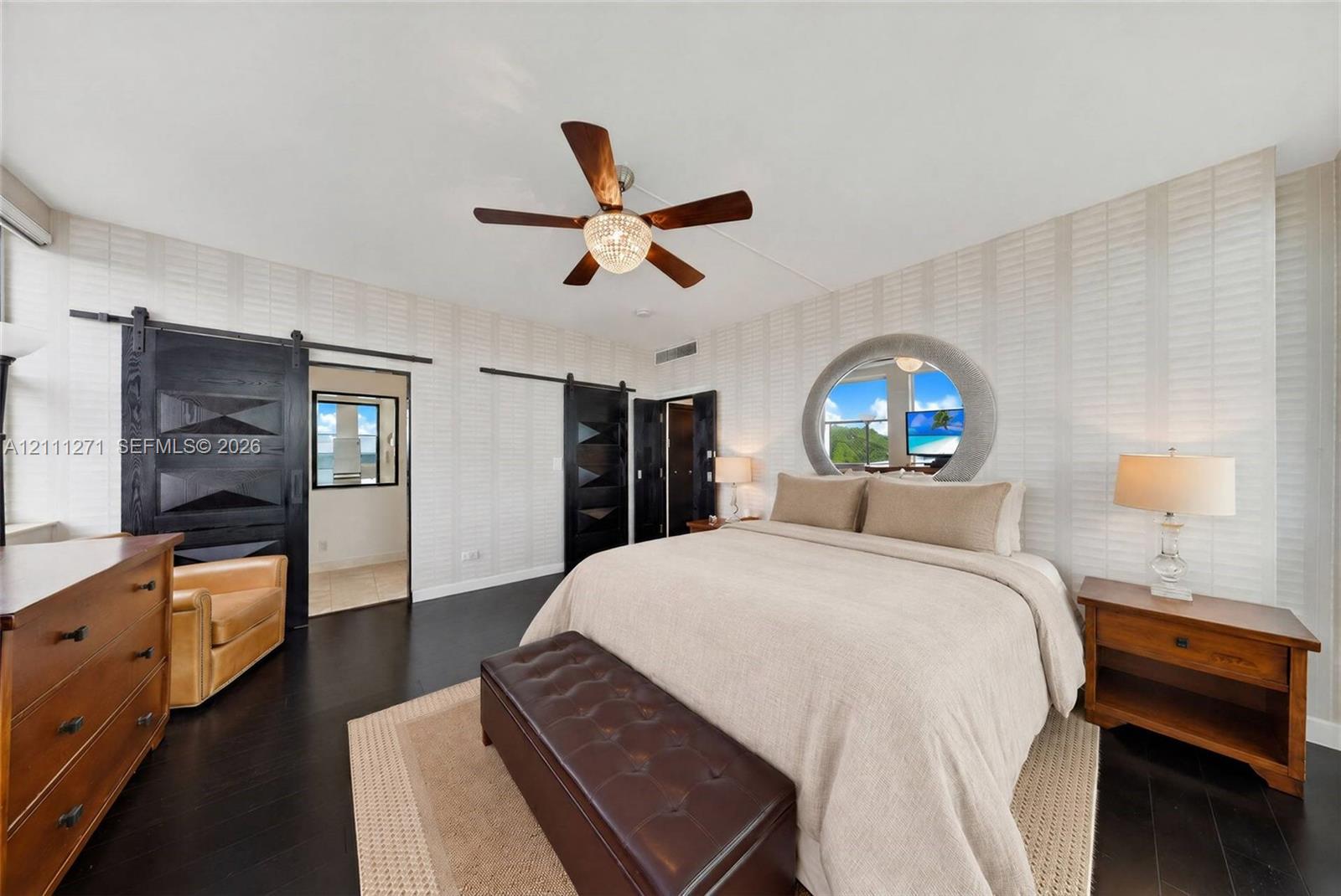 Dark wood-finish flooring, textured wallpaper, black paneled sliding barn doors, circular window with a coastal backdrop, and a ceiling fan with integrated lighting