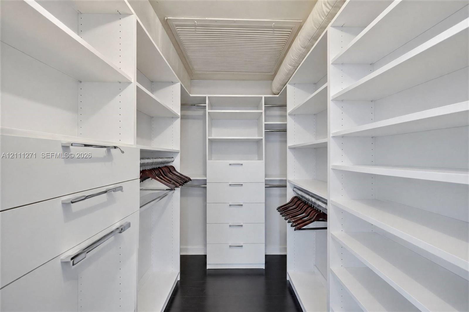 Walk-in closet featuring extensive built-in white shelving, multiple storage drawers with silver-tone hardware, and wood-finish flooring