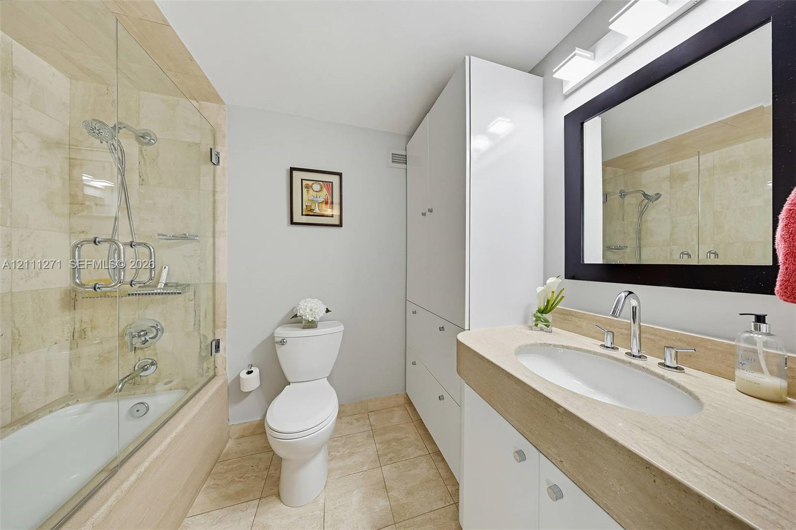 Bathroom featuring stone tile tub surround, frameless glass shower door, floor-to-ceiling built-in storage cabinetry, and vanity with an integrated stone countertop and oval undermount sink