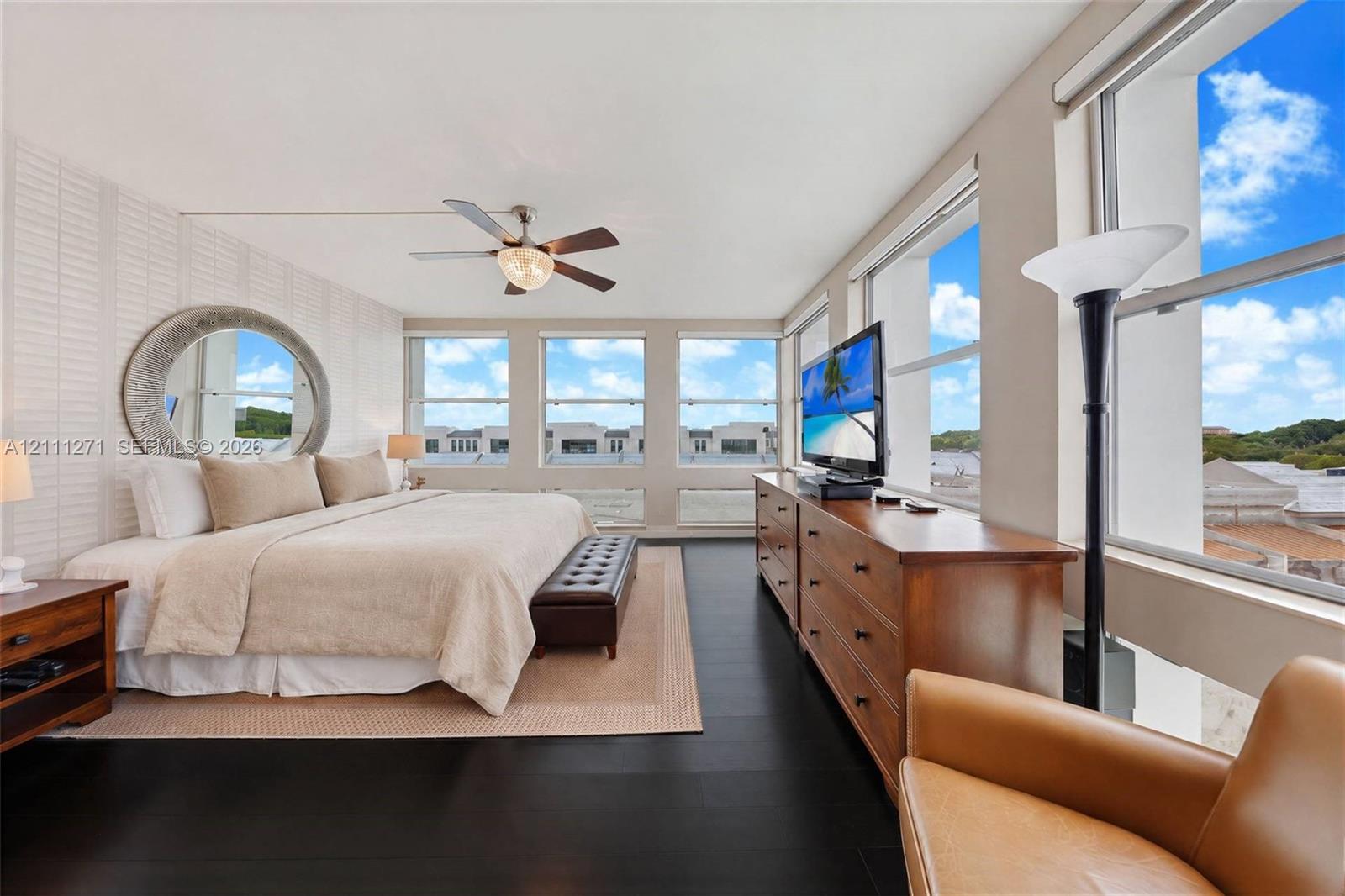 Spacious room featuring floor-to-ceiling windows, dark wood-finish flooring, a wall-mounted circular mirror, and a ceiling fan with integrated lighting