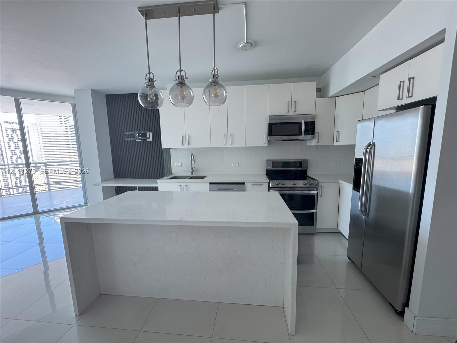 Kitchen featuring white flat-panel cabinetry, quartz waterfall island, stainless steel appliances, large-format tile flooring, and glass globe pendant lighting