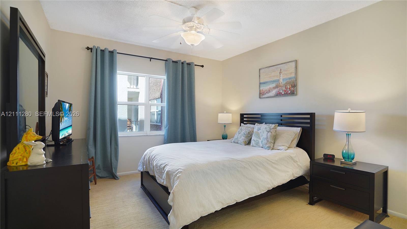 Neutral-toned interior room featuring wall-to-wall carpeting, a multi-pane window with dark metal curtain rod, and a central ceiling fan with integrated lighting
