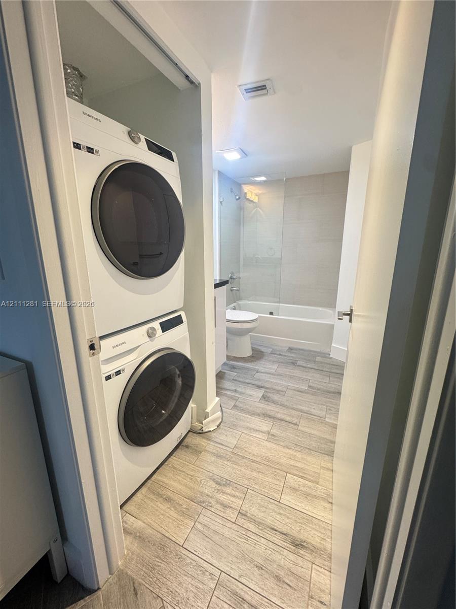 Stacked white appliance suite featuring a front-loading washer and dryer, complemented by wood-finish tile flooring, a bathtub with a glass panel, and neutral wall tile surrounds