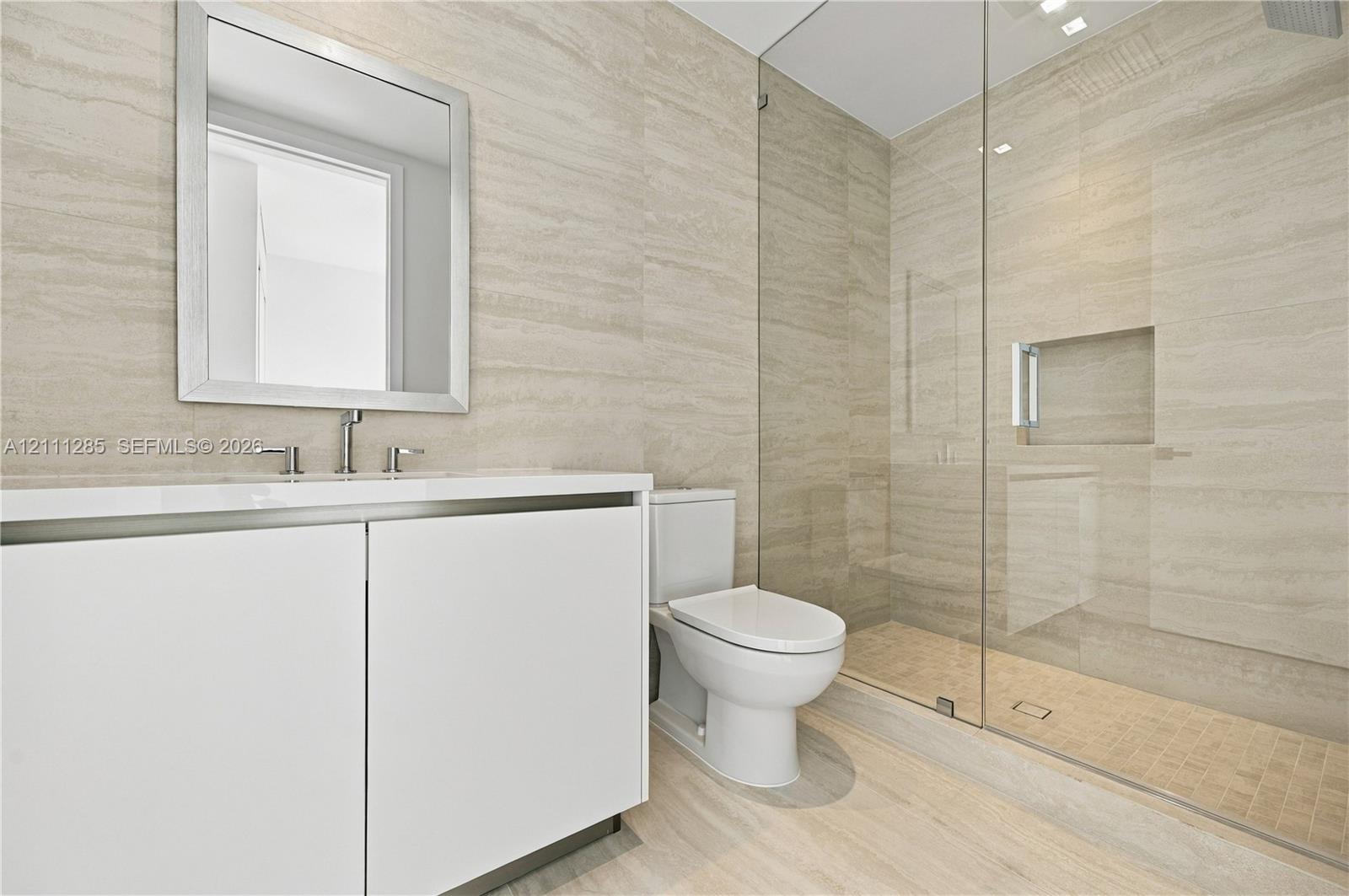 Minimalist vanity featuring a smooth-finish white cabinet and rectangular sink with metal fixtures, complemented by wall-to-wall linear-patterned tile, a seamless glass-enclosed shower with a built-in recessed niche, and mosaic floor tiling