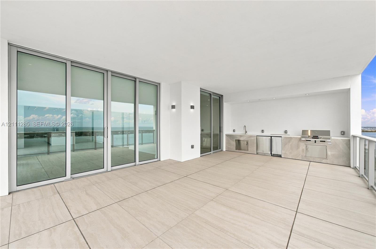 Expansive covered terrace featuring large-format tile flooring, a built-in outdoor kitchen with stainless steel appliances, stone-finish cabinetry, floor-to-ceiling glass sliding doors, and recessed lighting