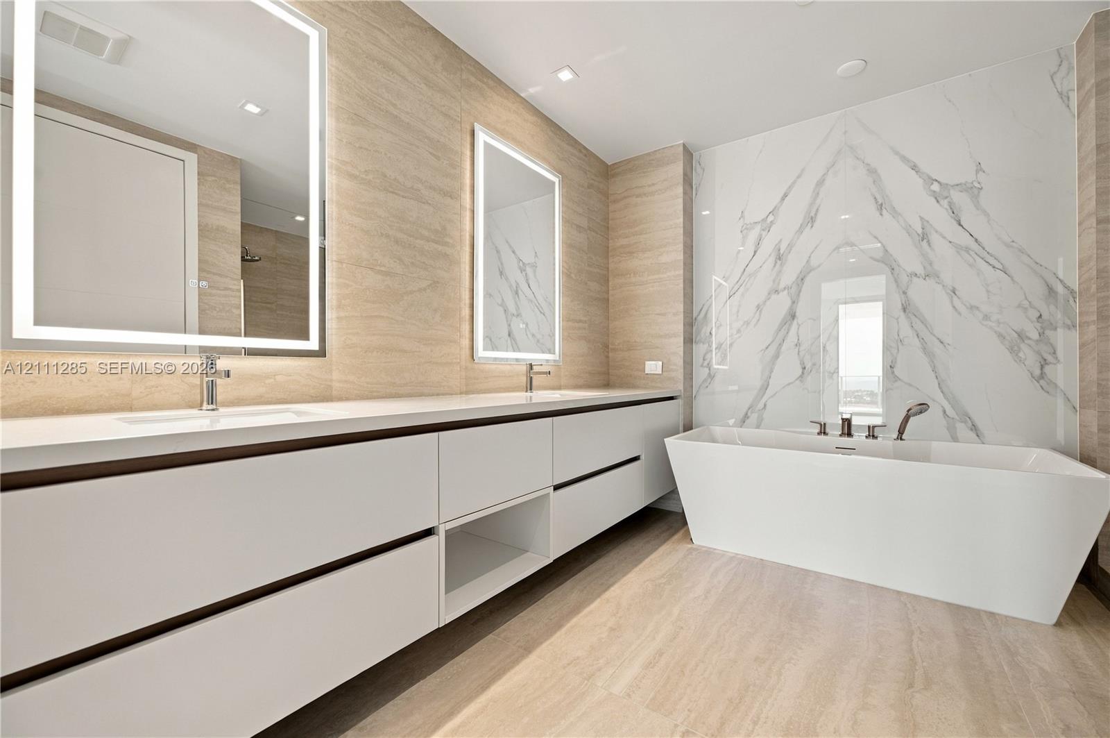 Dual-sink vanity with white cabinetry and countertop, backlit rectangular mirrors, freestanding soaking tub, and large-format tile surround