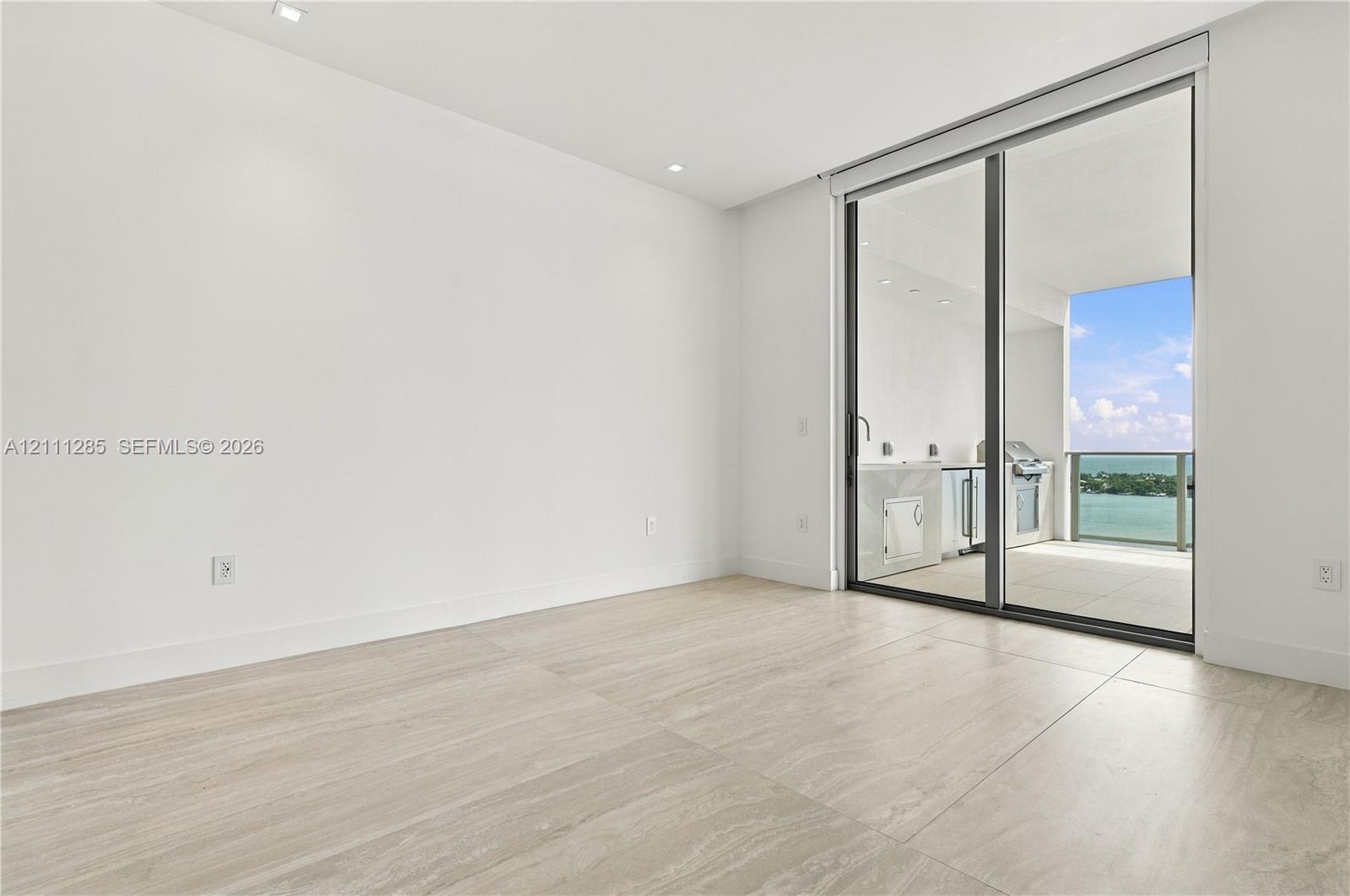 Large-format tile flooring, recessed ceiling lighting, and floor-to-ceiling sliding glass doors opening to a covered balcony with built-in cabinetry