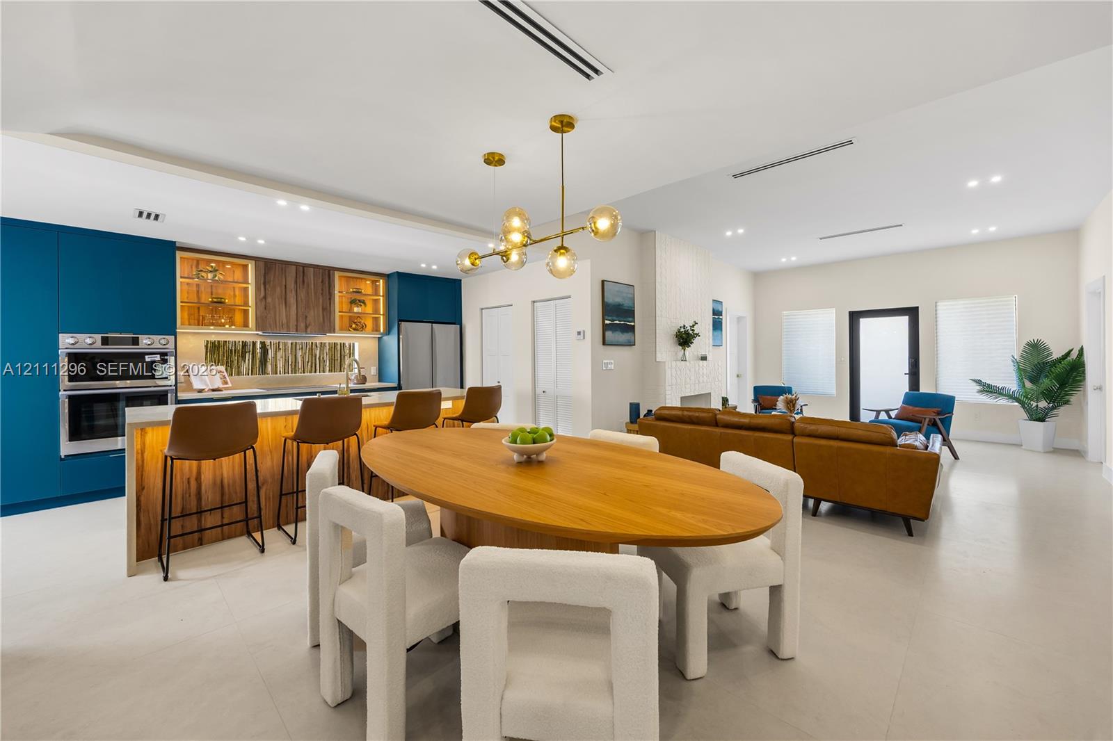Modern kitchen featuring deep blue flat-panel cabinetry, integrated stainless steel double wall ovens, wood-finish accent shelving, a minimalist multi-globe pendant light, and neutral tile flooring