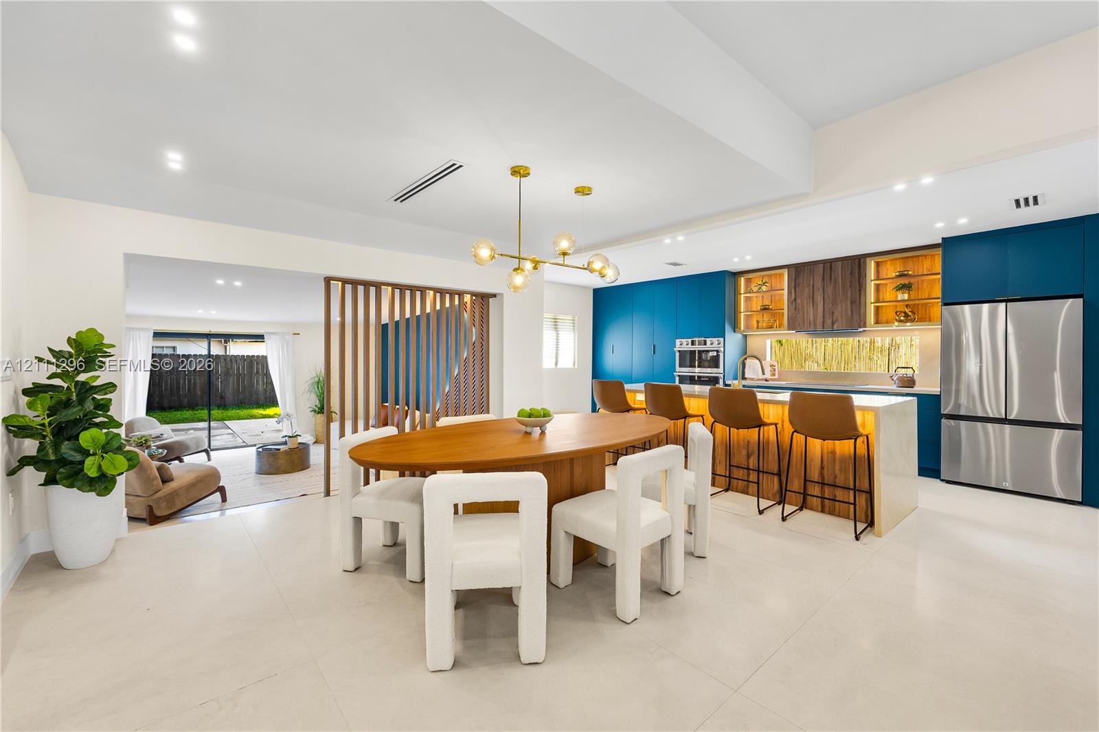 Open-concept kitchen and dining area featuring a central island with integrated stone countertop, modern blue cabinetry, wooden slat room divider, and brushed gold chandelier lighting Open-concept kitchen and dining area featuring a central island with integrated stone countertop, modern blue cabinetry, wooden slat room divider, and brushed gold chandelier lighting