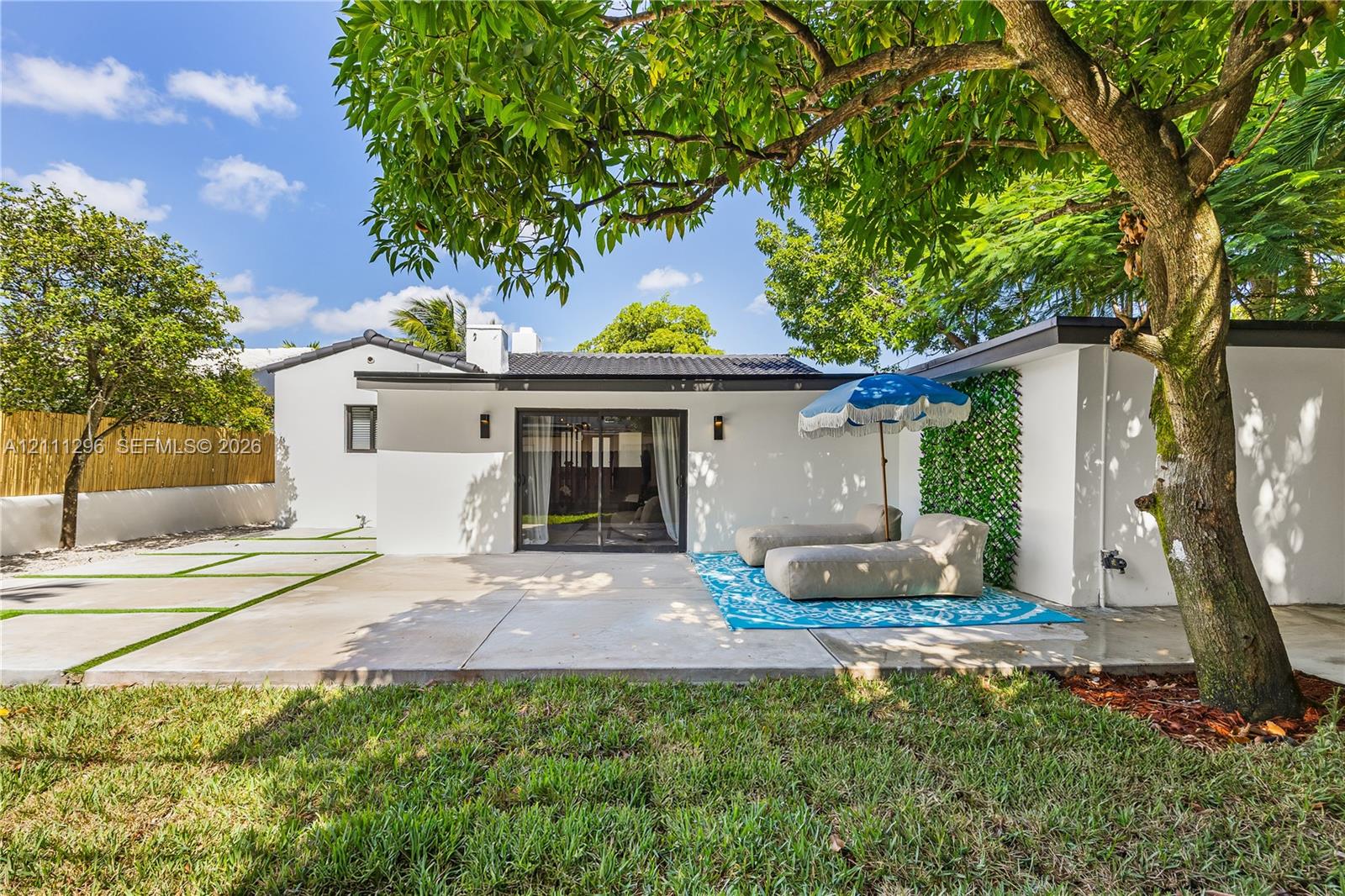 Rear exterior featuring a concrete patio, sliding glass doors, mature trees, a bamboo privacy screen, and exterior wall-mounted lighting