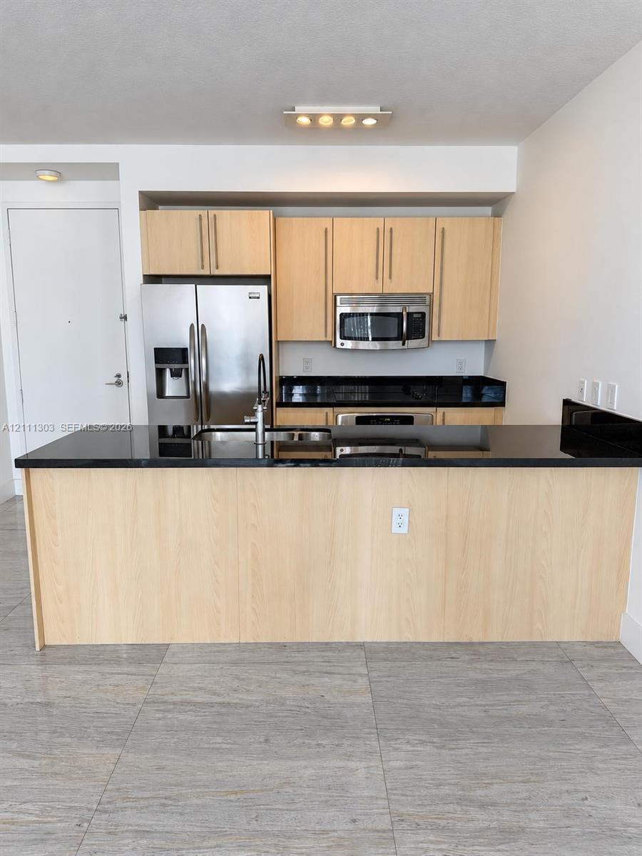 Kitchen featuring a central island with a solid surface countertop, light wood-finish cabinetry, stainless steel appliances, and large-format tile flooring Kitchen featuring a central island with a solid surface countertop, light wood-finish cabinetry, stainless steel appliances, and large-format tile flooring