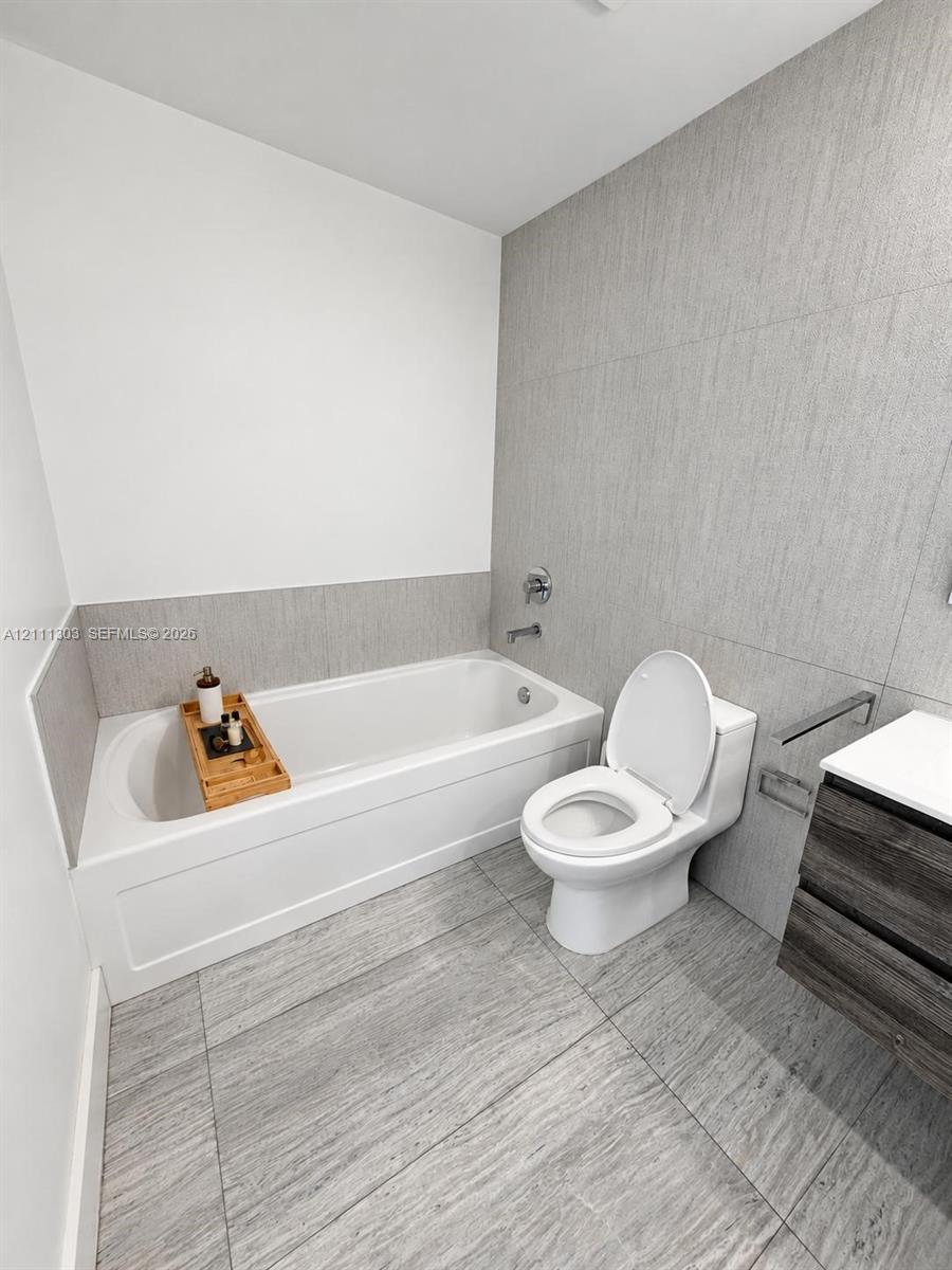 Bathroom featuring a deep soaking tub, large-format textured tile flooring, full-height wall tiling, and a floating vanity with a stone countertop