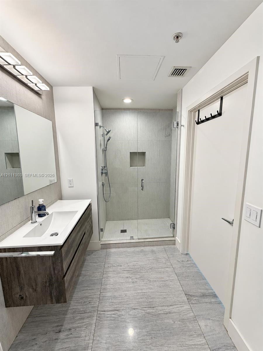Floating wood-finish vanity with integrated sink, stone-composite countertop, glass-enclosed shower with tiled surround, and large-format tile flooring