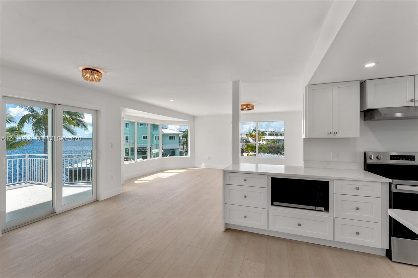 Open-concept kitchen and living area featuring wood-finish flooring, white shaker cabinetry, stainless steel appliances, large bay windows, and sliding glass doors to a waterfront balcony