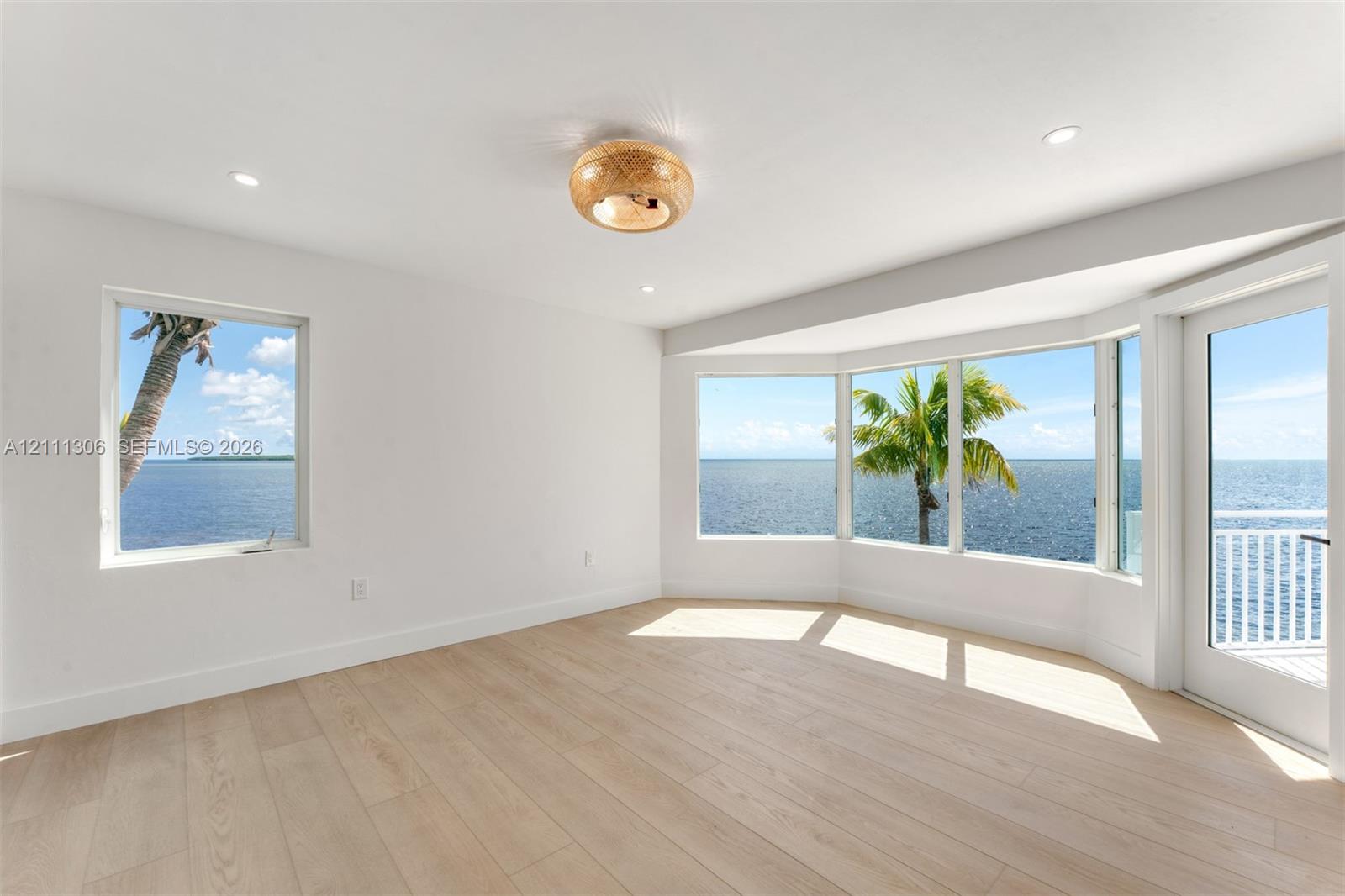 Light-filled room featuring wood-finish flooring, a bay window, recessed lighting, and a glass-paneled door opening to an exterior balcony with water and palm tree scenery