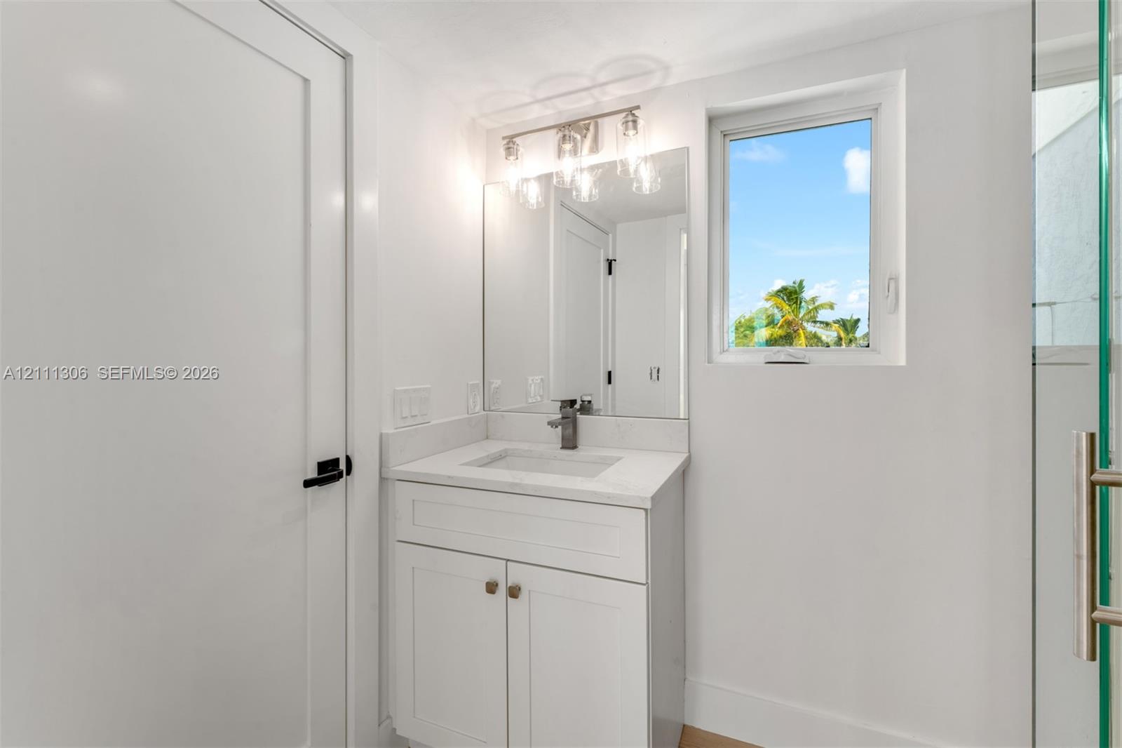 White shaker-style vanity with a solid surface countertop, integrated rectangular sink, three-light fixture with glass shades, wall-mounted mirror, and glass-enclosed shower