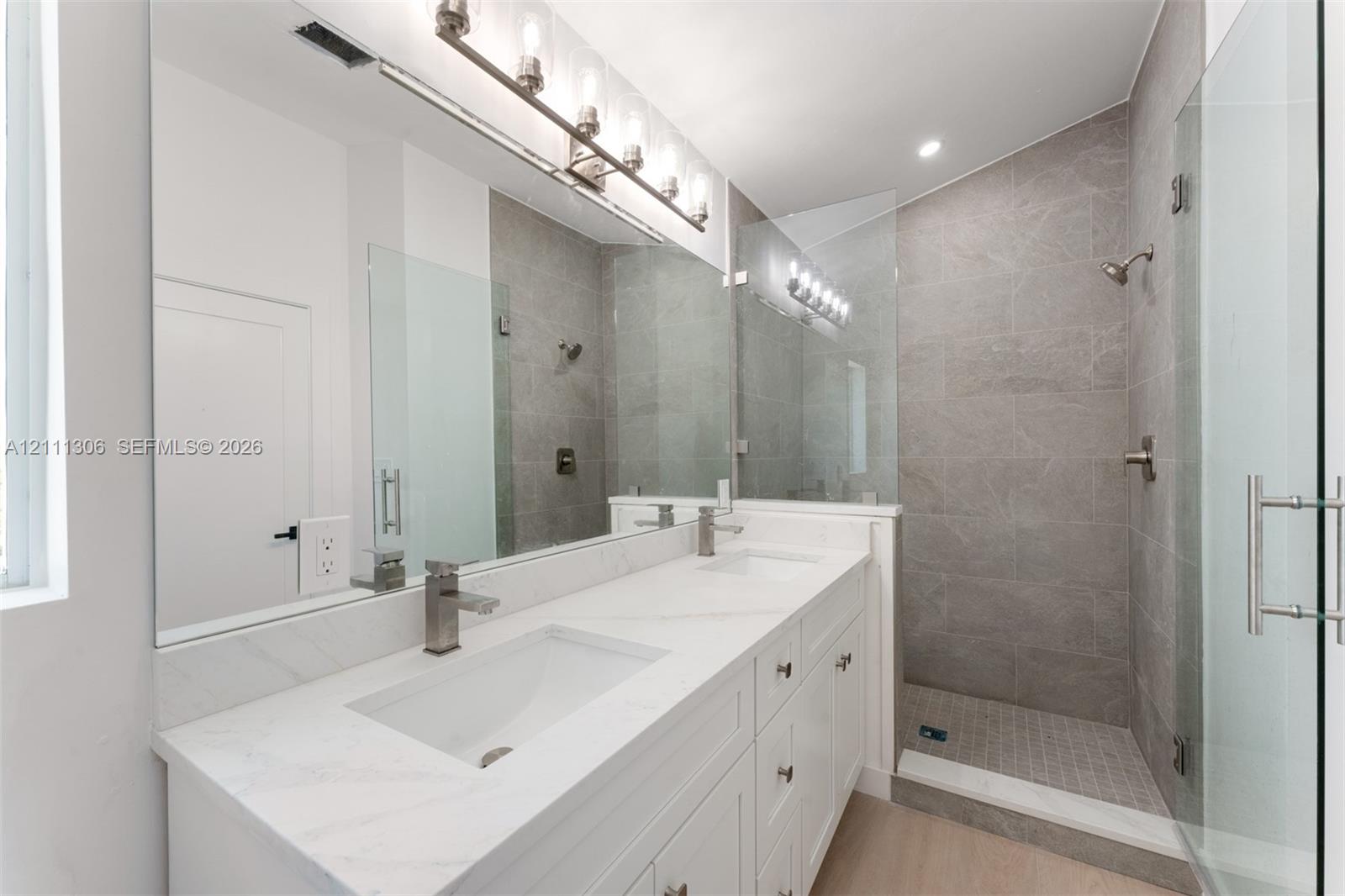 Double-vanity with stone countertop, walk-in shower featuring tile surround and glass enclosure, large wall-mounted mirror, and multi-bulb vanity lighting