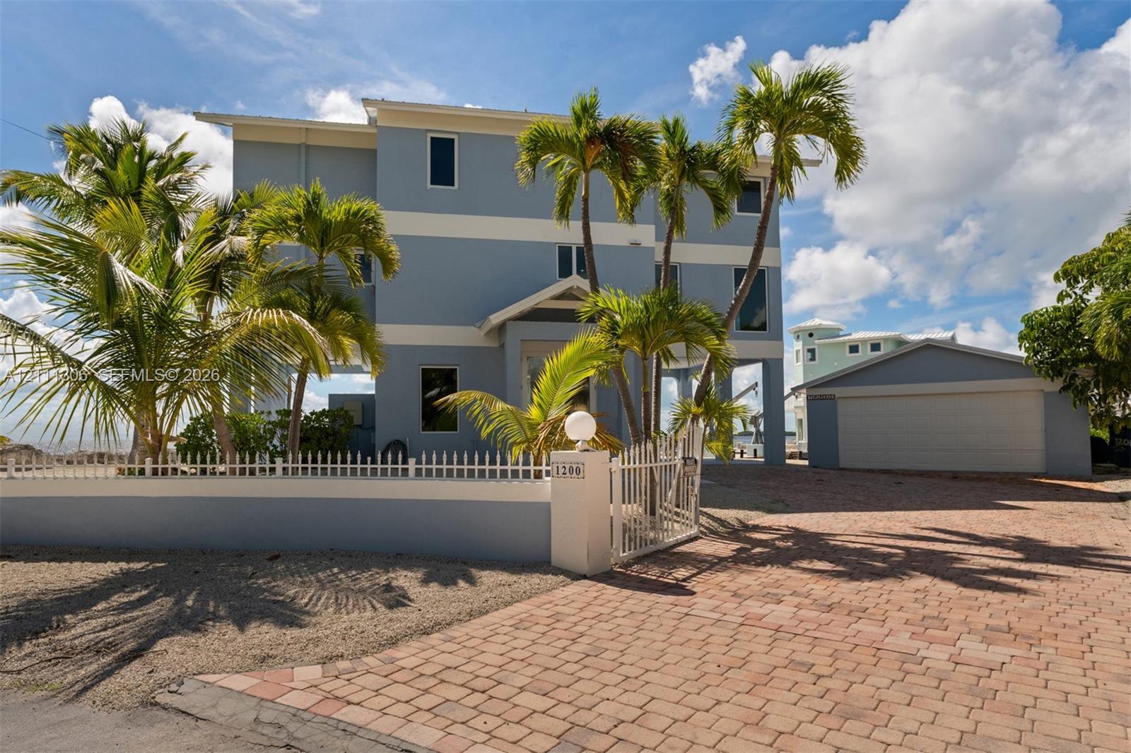 Three-story stilt-constructed exterior featuring a light blue facade, paver driveway, detached two-car garage, and perimeter masonry fencing