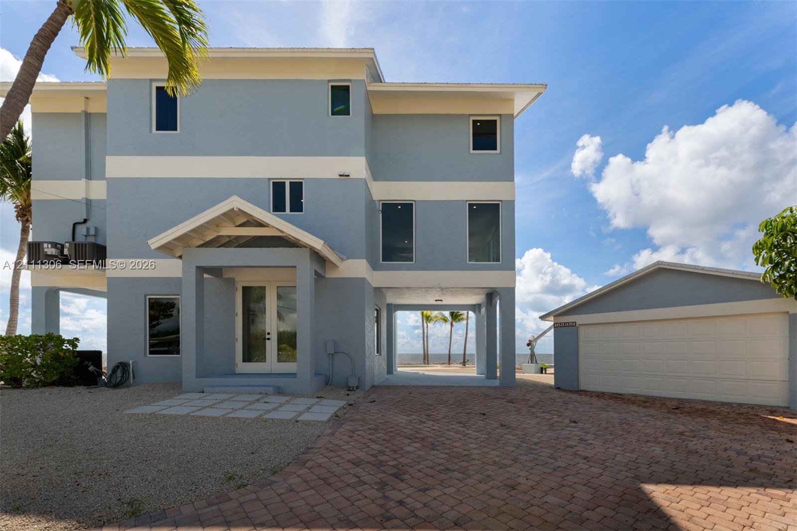 Three-story elevated residence featuring a paver driveway, detached garage, covered entryway with double doors, and reinforced concrete pilings