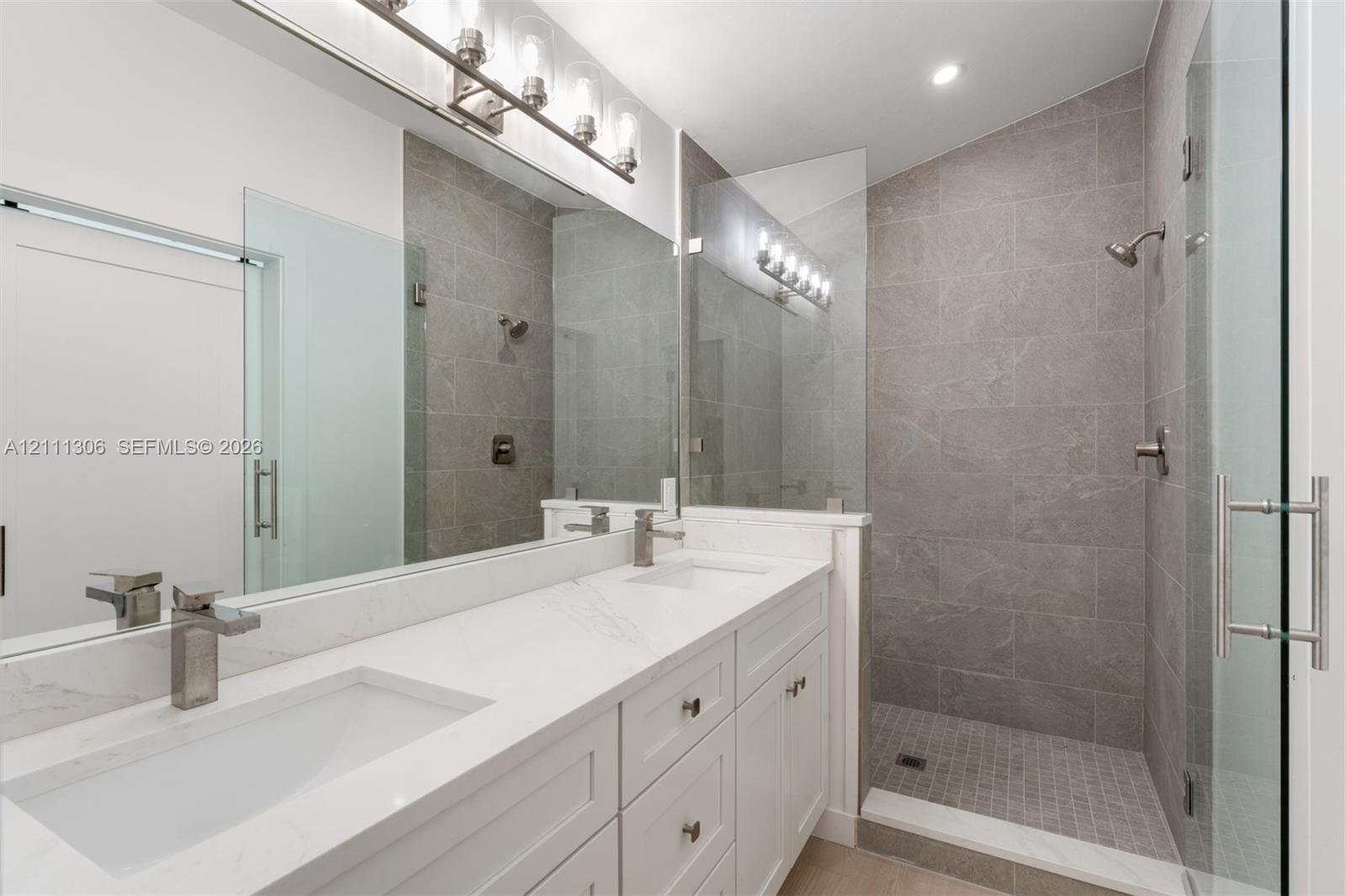 Dual-sink vanity with stone-finish countertops and chrome fixtures, paired with a walk-in shower featuring large-format tile surround, mosaic floor tiling, and a glass enclosure