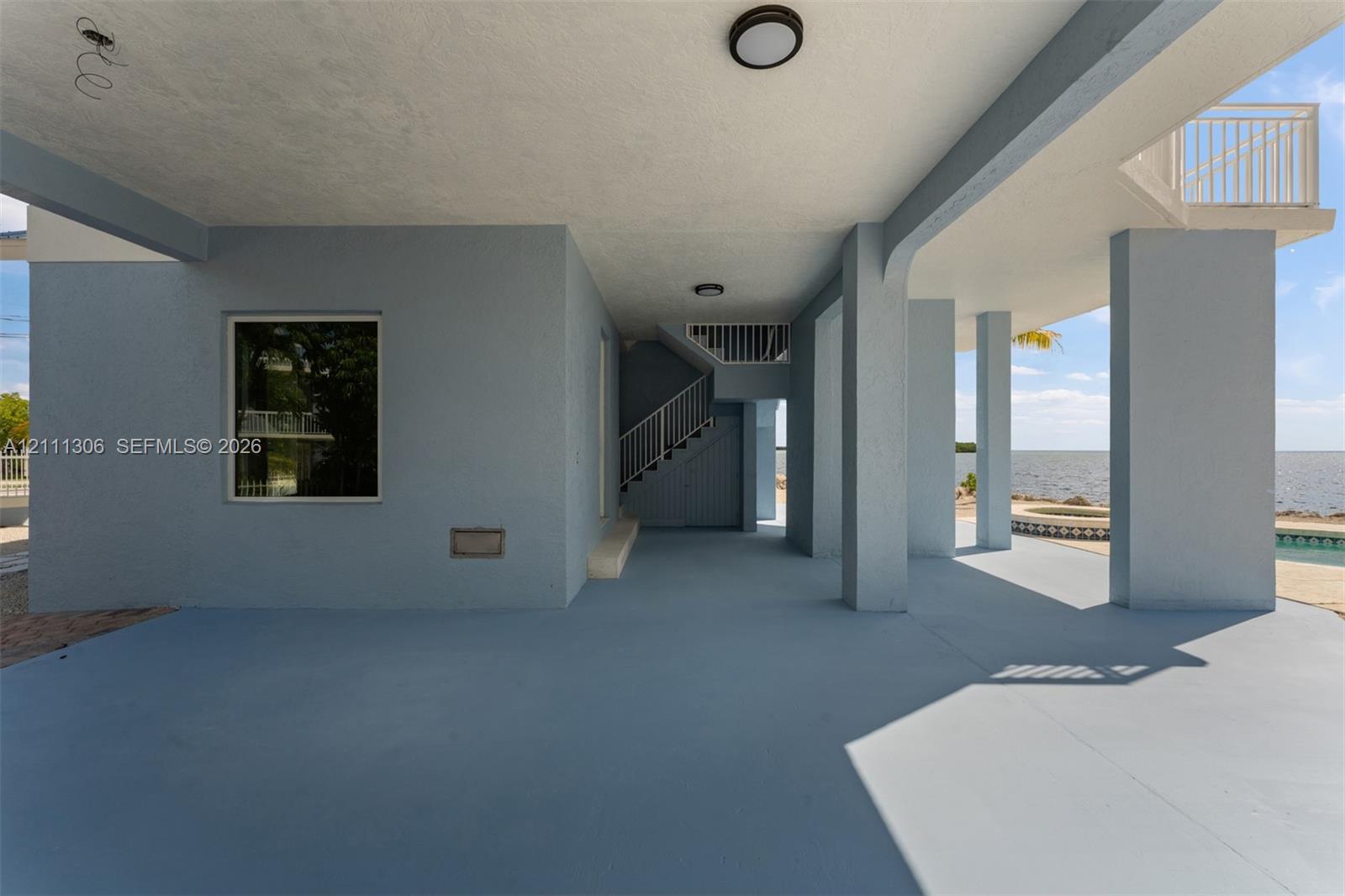 Covered ground-level patio featuring structural concrete columns, staircase with metal railings, flush-mount lighting, and exterior access to a pool area with water backdrop