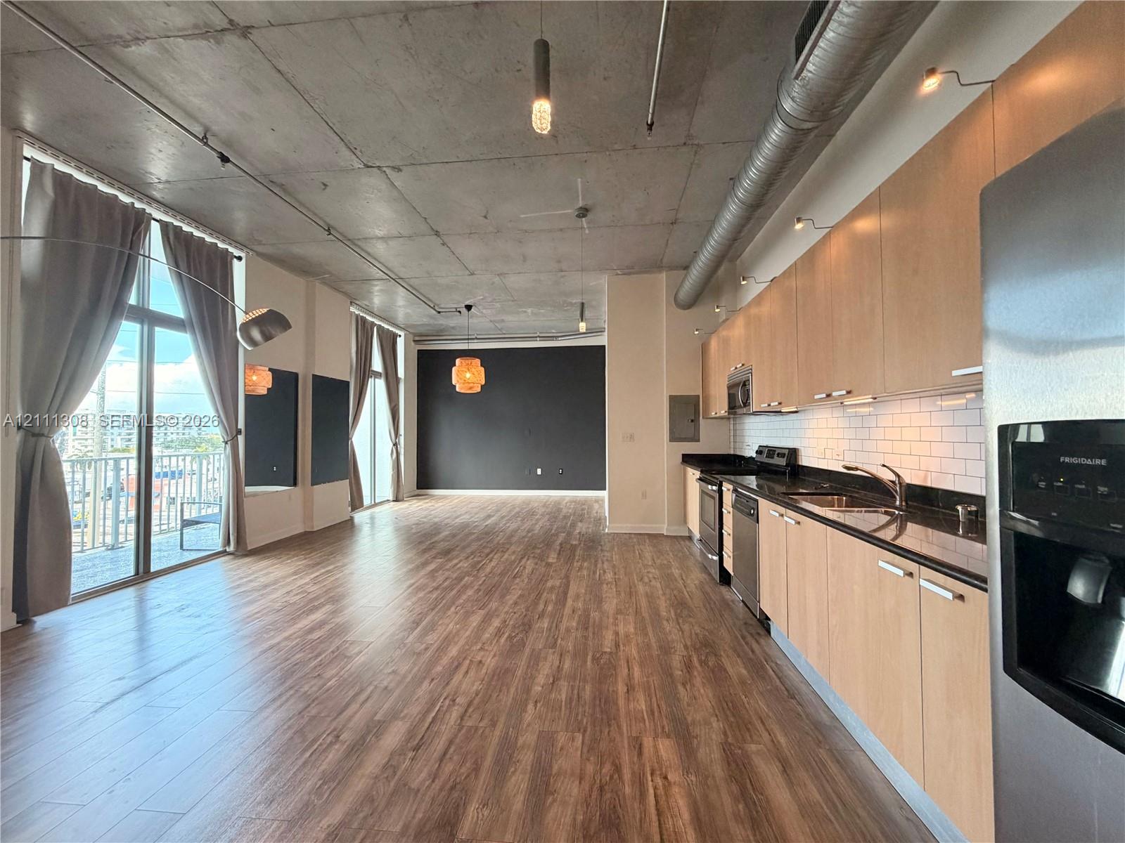 Industrial-style interior featuring exposed concrete ceilings, wood-finish flooring, floor-to-ceiling windows, and a kitchen equipped with flat-panel cabinetry and stone countertops Industrial-style interior featuring exposed concrete ceilings, wood-finish flooring, floor-to-ceiling windows, and a kitchen equipped with flat-panel cabinetry and stone countertops