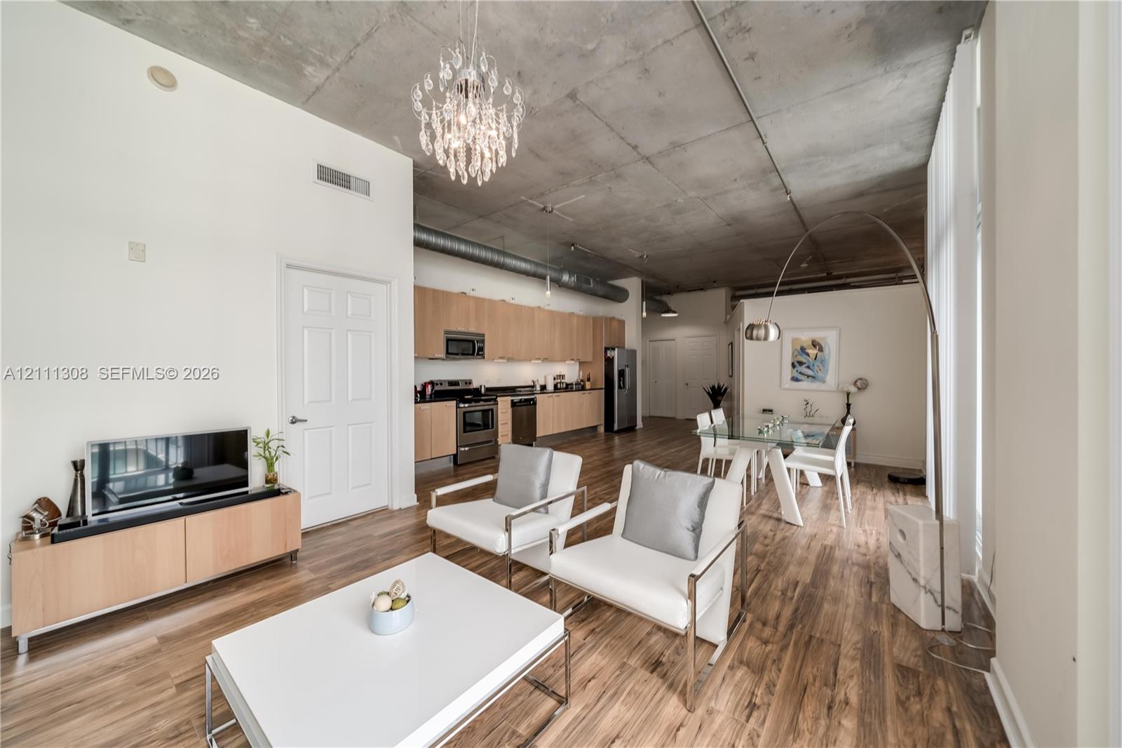 Open-concept industrial-style interior featuring exposed concrete ceilings, wood-finish flooring, custom flat-panel cabinetry, stainless steel kitchen appliances, and a crystal chandelier Open-concept industrial-style interior featuring exposed concrete ceilings, wood-finish flooring, custom flat-panel cabinetry, stainless steel kitchen appliances, and a crystal chandelier