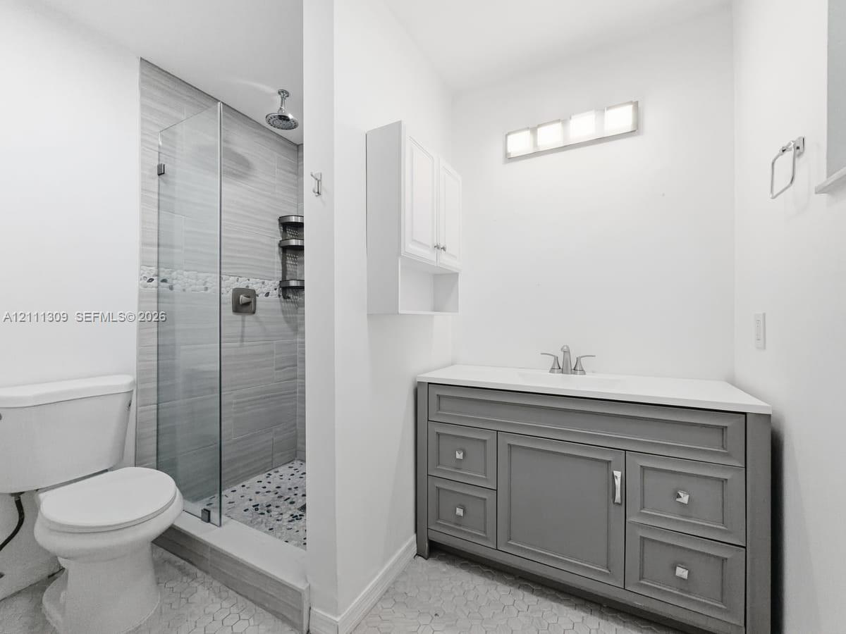 Bathroom featuring a grey vanity with a solid surface countertop, glass-enclosed walk-in shower with horizontal tile walls, pebble-style floor tiling, and wall-mounted cabinetry