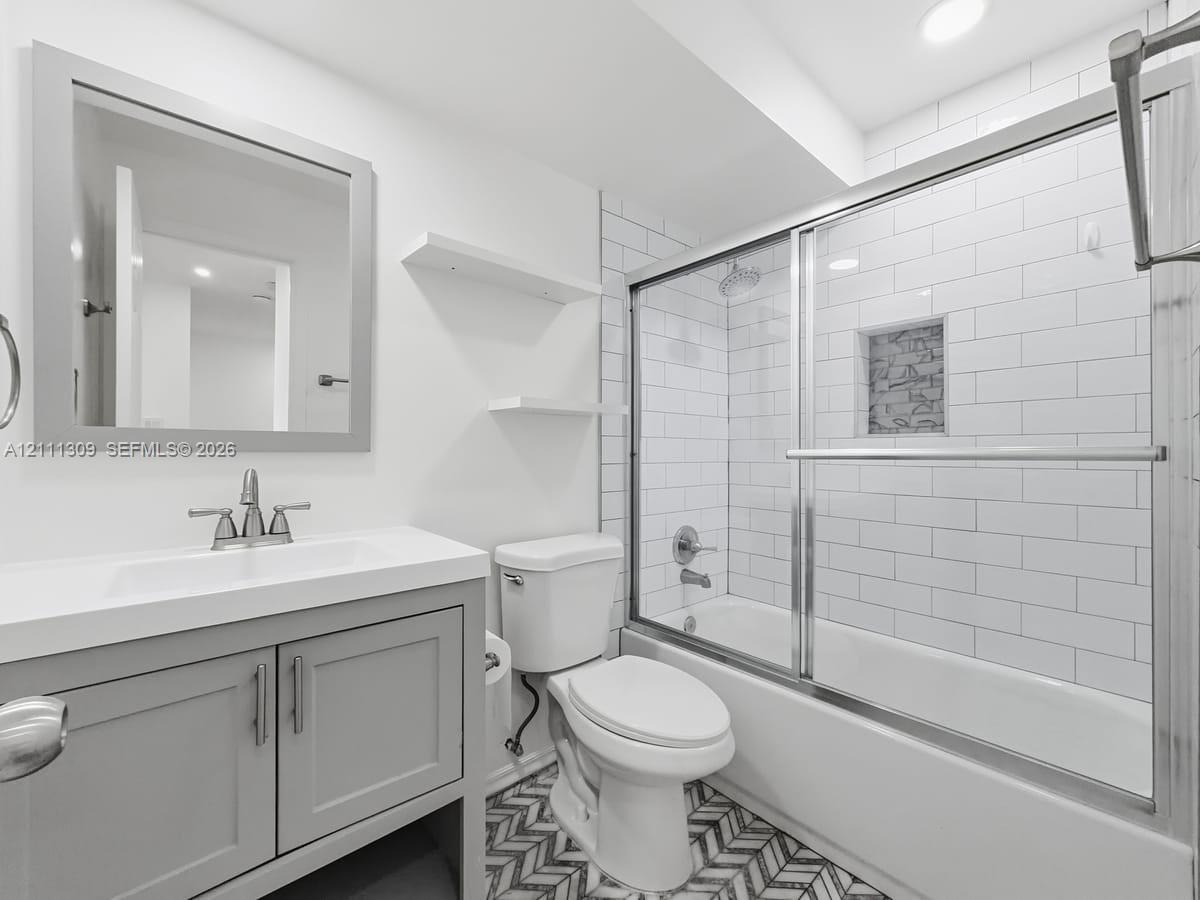 Grey vanity cabinet with integrated sink and countertop, white subway tile shower surround with built-in wall niche, glass sliding shower door, and patterned tile flooring