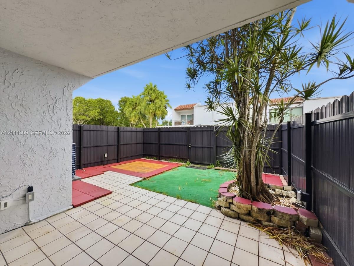 Covered patio with tiled flooring, enclosed by dark vertical-panel fencing, featuring a raised stone planter border and mature landscaping