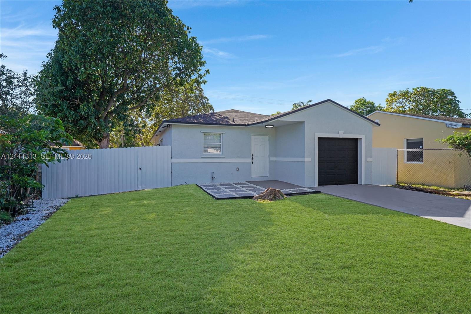 Single-story residence featuring a light-toned stucco exterior, a concrete driveway, an attached garage with a dark-finished door, and a privacy fence