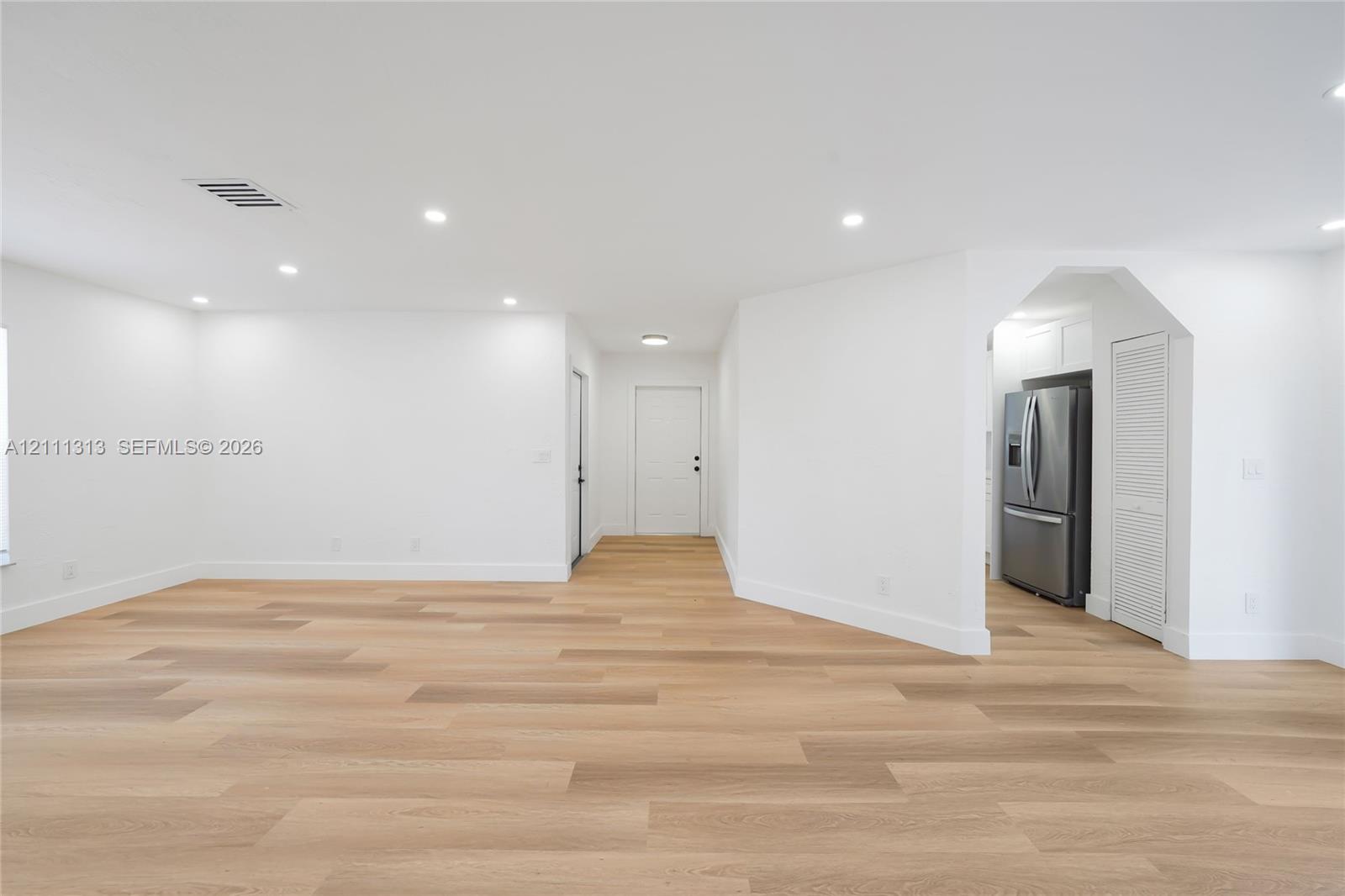 Open-concept interior featuring light wood-finish flooring, recessed lighting, smooth white walls, and a stainless steel refrigerator